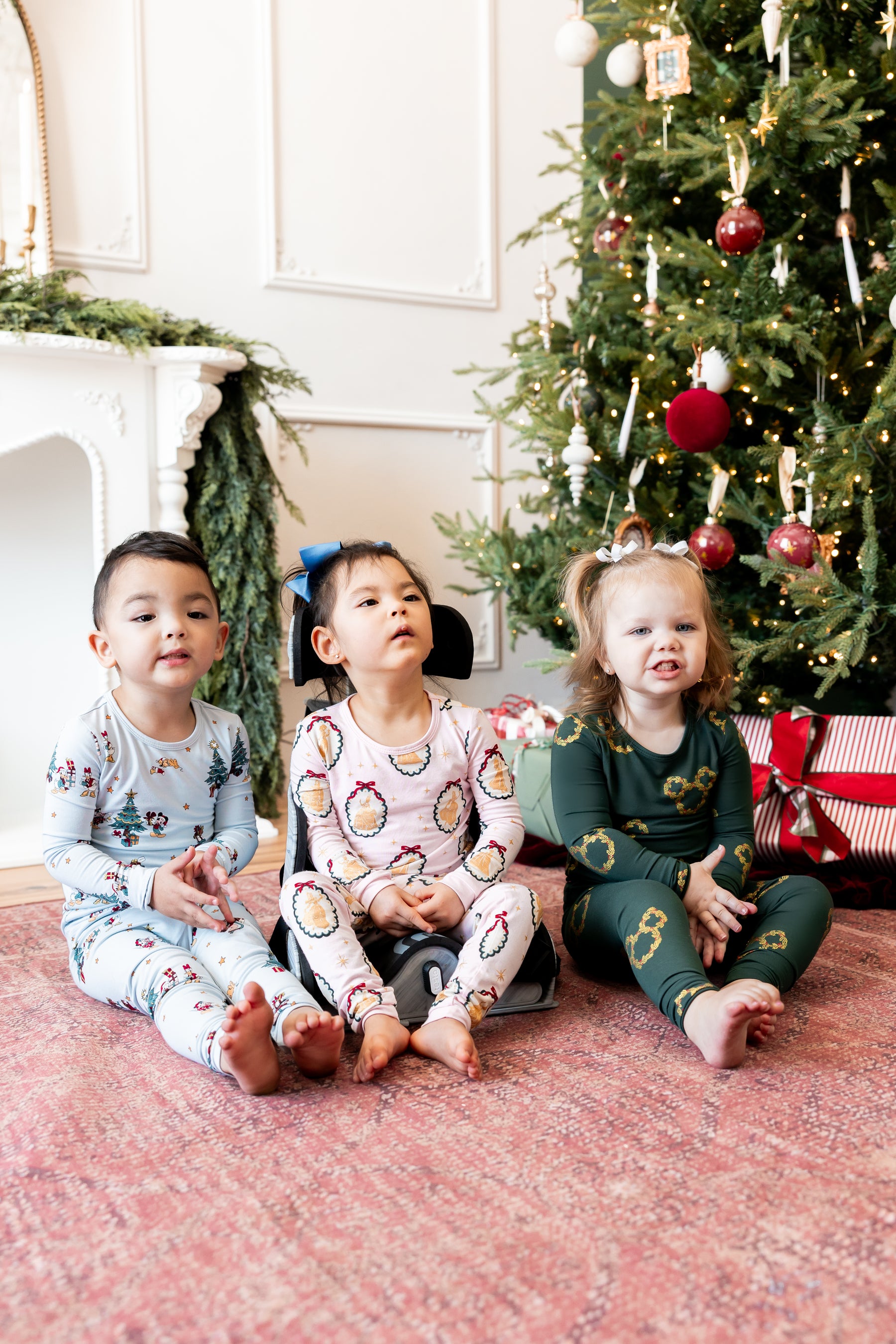 Three kids wearing Long Sleeve Pajamas in Mickey & Friends Christmas print, Mickey Christmas Wreath and Princess portrait sitting together beside a Christmas tree