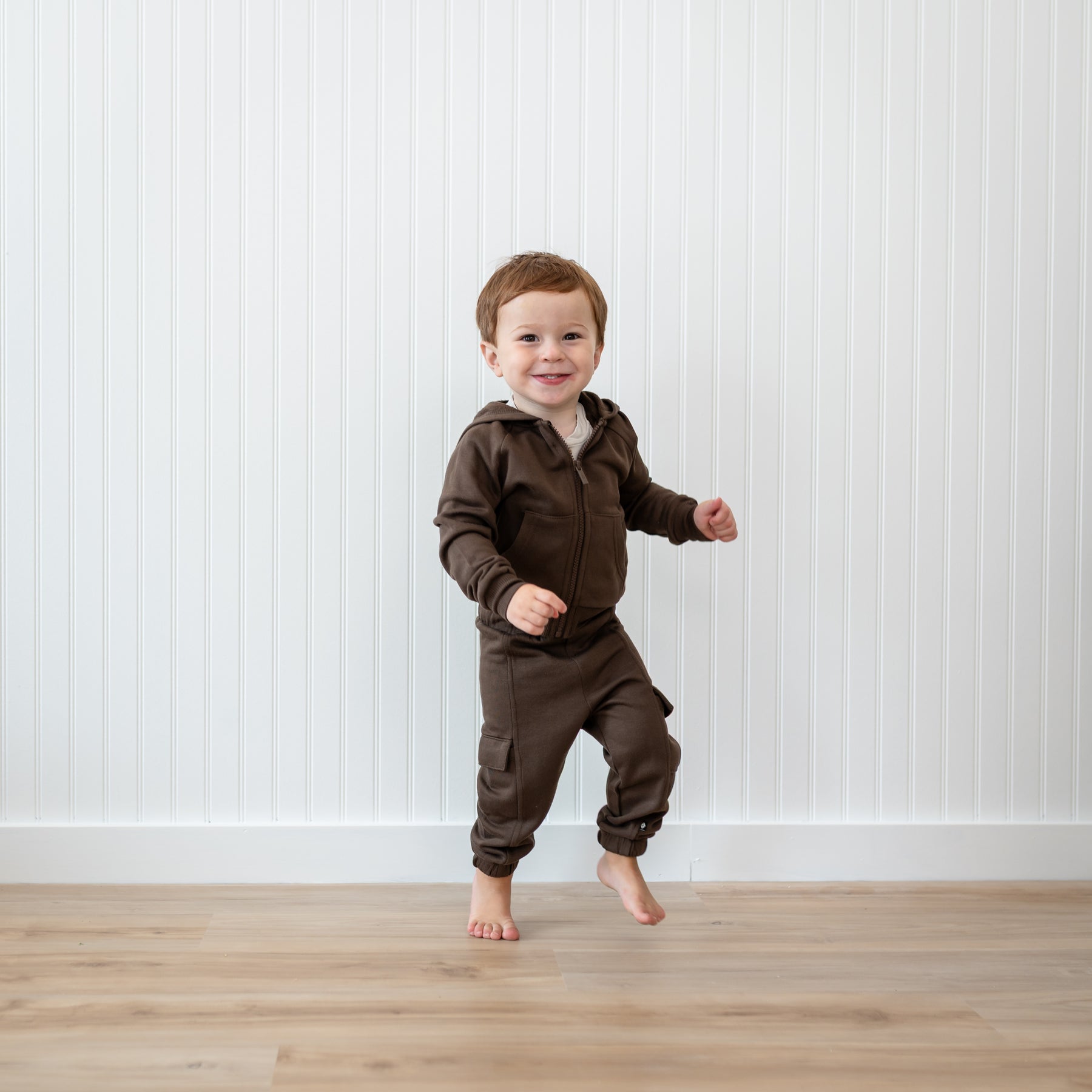 Young boy jumping in front of a white paneled wall wearing the Fleece Zip Up Hoodie in Espresso and matching fleece cargo pant