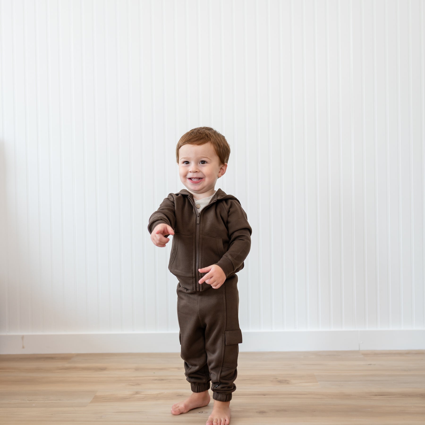 Young boy standing in front of a white paneled wall wearing the Fleece Zip Up Hoodie in Espresso and matching fleece cargo pants pointing forwards