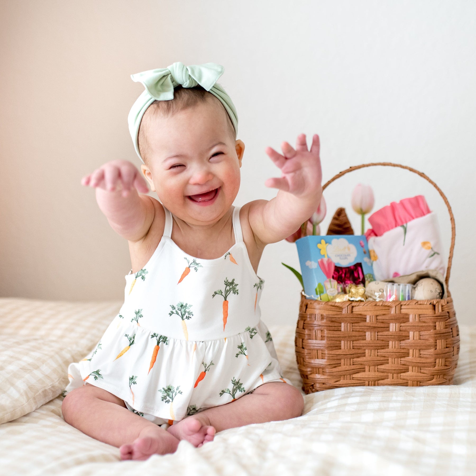 Smiling toddler sitting on a bed wearing the Spaghetti Strap Ruffle Romper in Carrot beside an easter basket