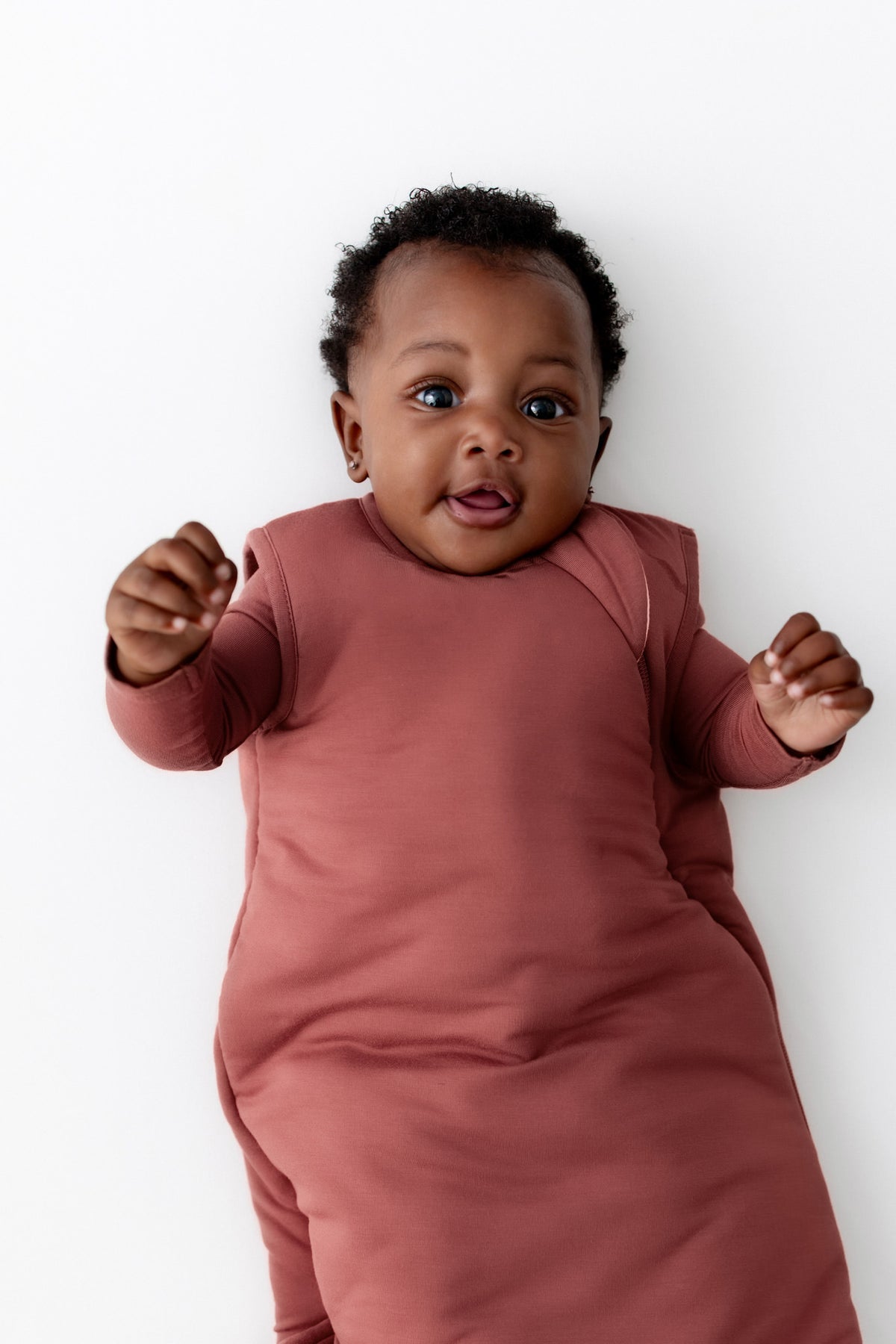 A baby lying on their back wearing a terracotta wearable blanket against a clean white background.