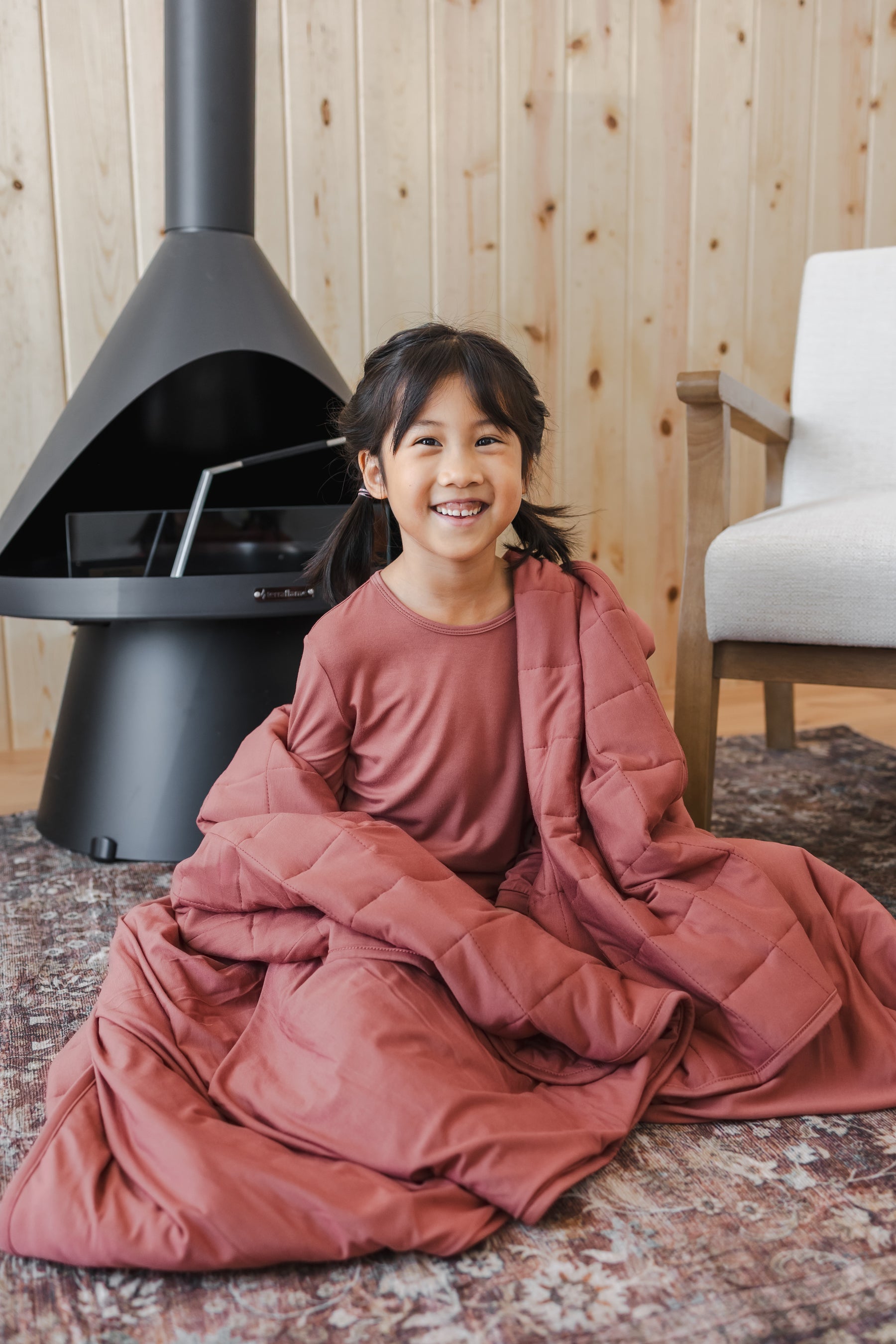 Girl sitting on floor wrapped in Youth Blanket in Ember 2.5 wearing a similar color shirt