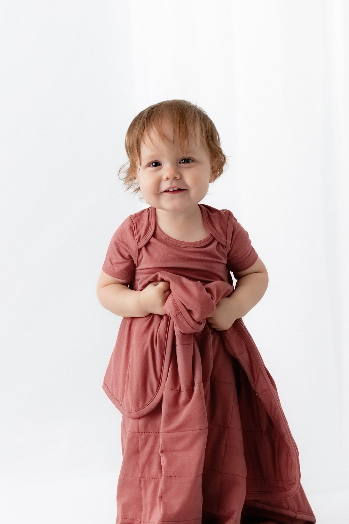 Baby holding Baby Blanket in Ember while wearing a similar color bodysuit dress on a white background