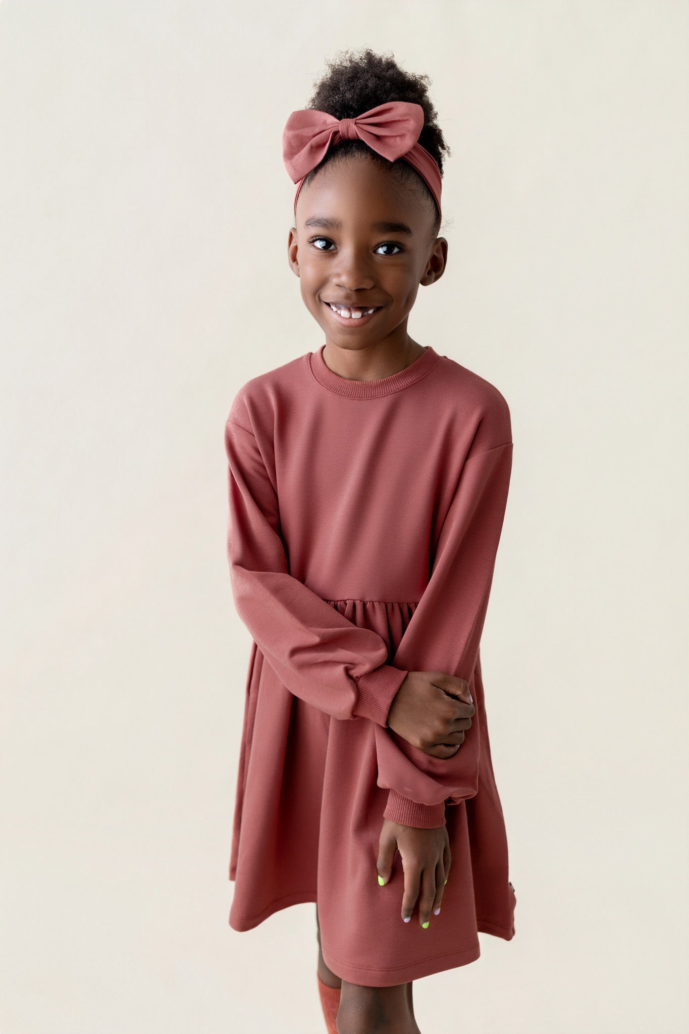 Girl wearing Long Sleeve Skater Dress in Ember color with knotted bow headband on a beige background