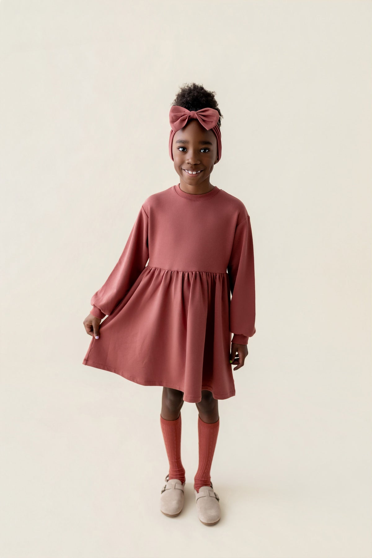 Girl posing in Long Sleeve Skater Dress in Ember color with knotted bow headband on a beige background