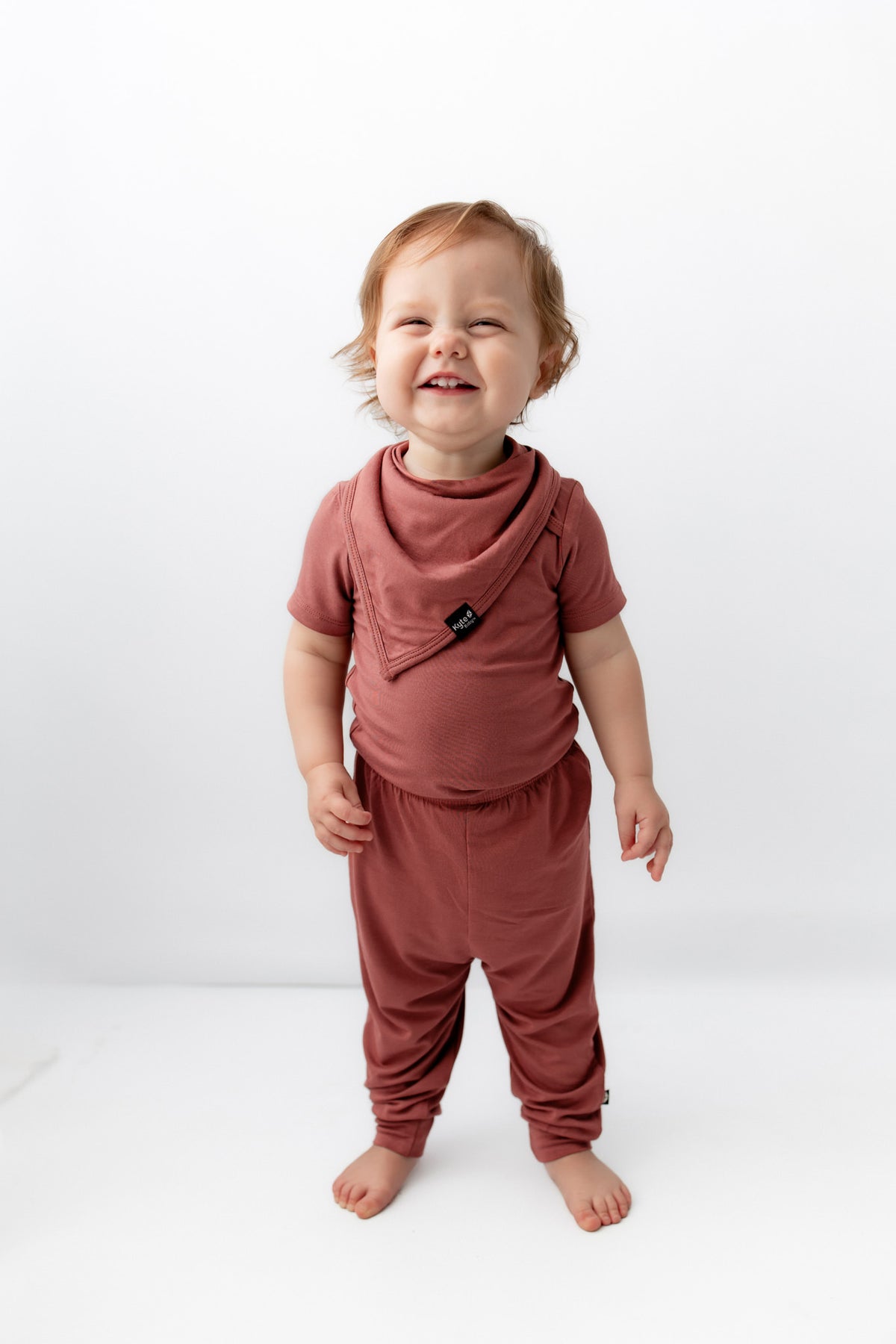Child smiling while wearing Bib in Ember over similar color shirt and pant on a white background