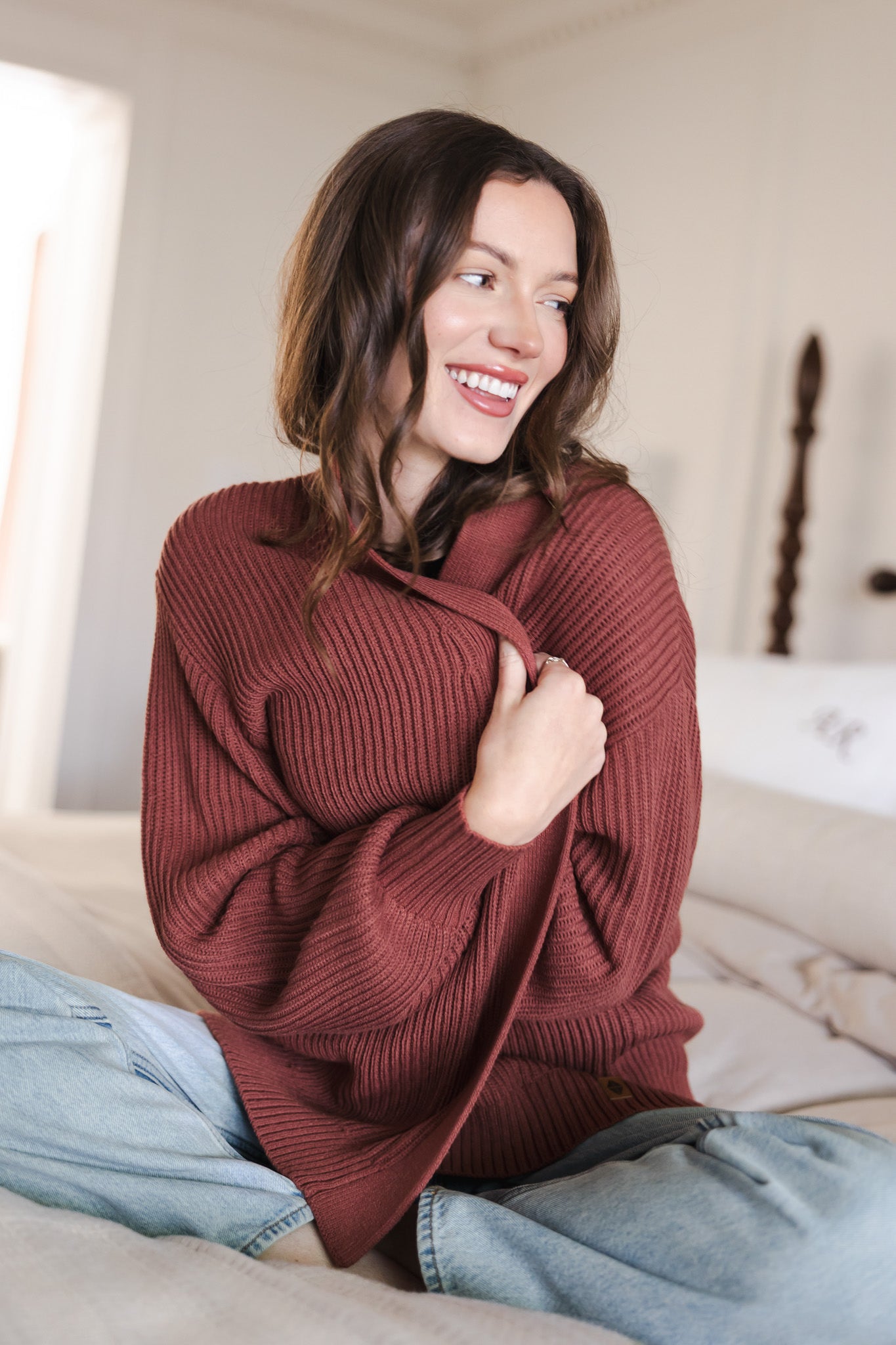 Modal sitting on beige bed wearing Chunky knit oversized cardigan in ember over blue jeans