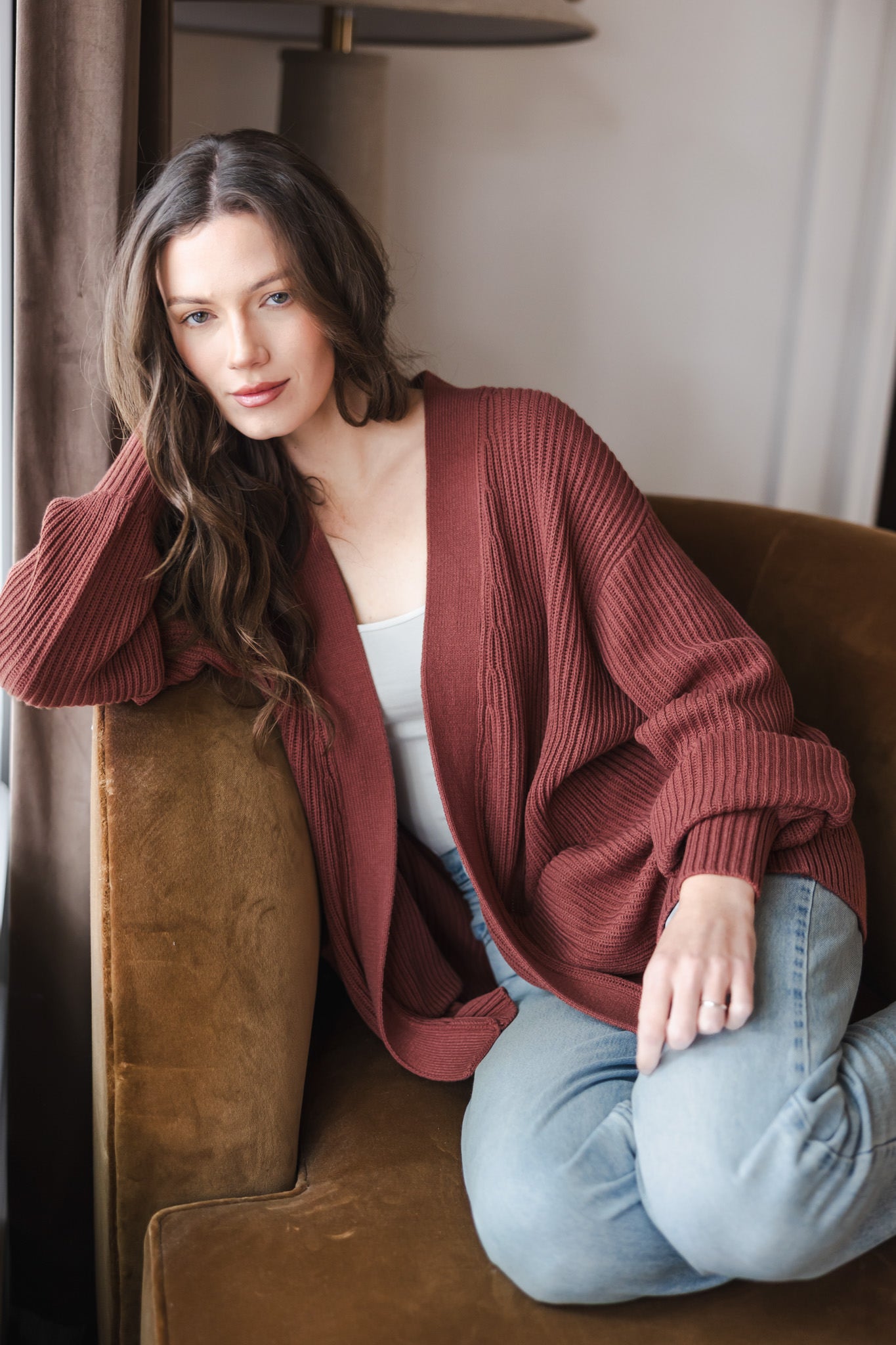 Modal posing on brown sofa wearing Chunky knit oversized cardigan in ember over white t-shirt and blue jeans