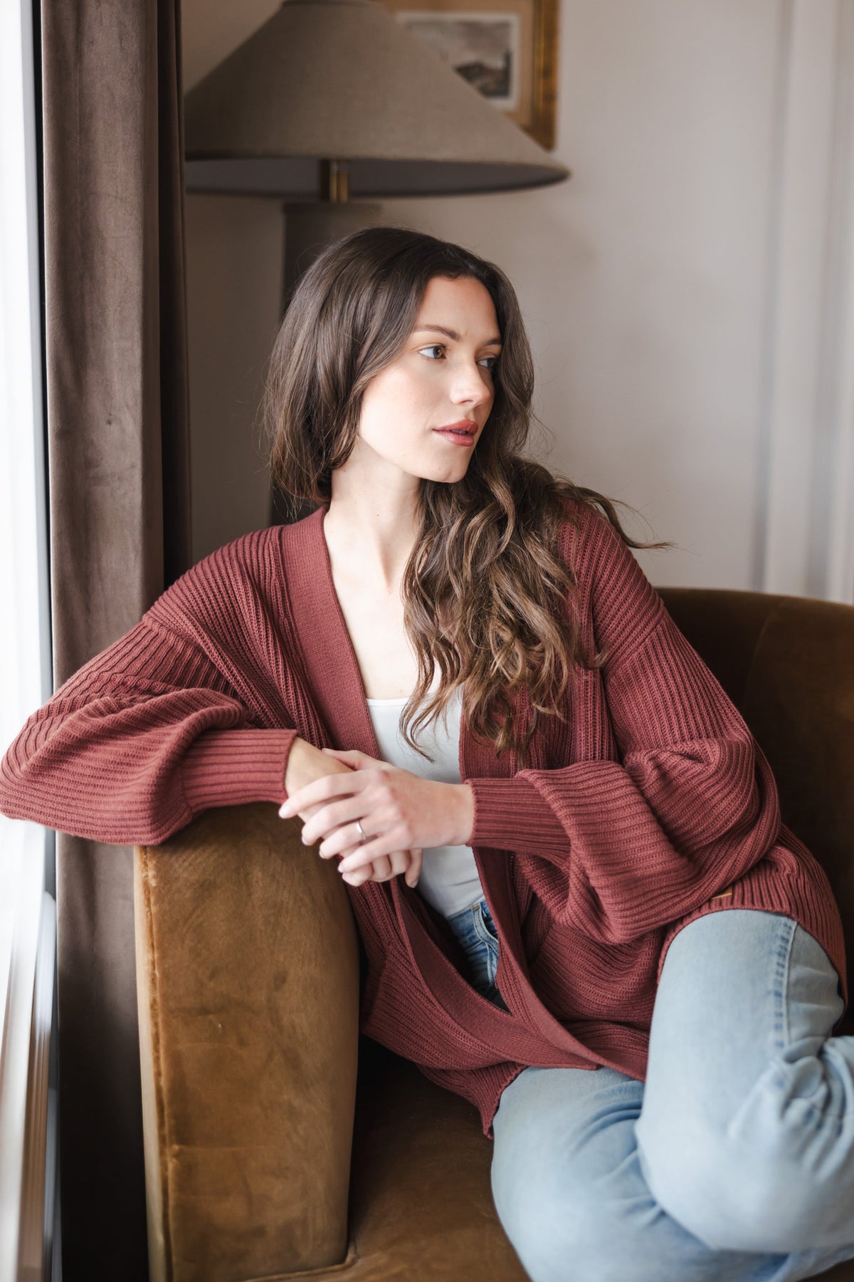 Modal sitting on brown sofa wearing Chunky knit oversized cardigan in ember over white t-shirt and blue jeans