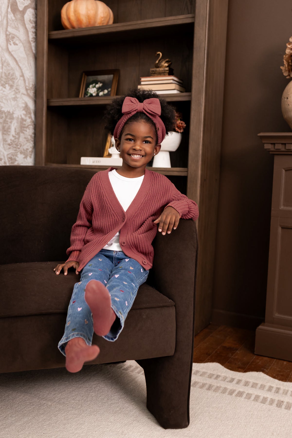 Girl sitting on a sofa wearing Chunky Knit Oversized Cardigan in Ember over White T-shirt and blue jeans