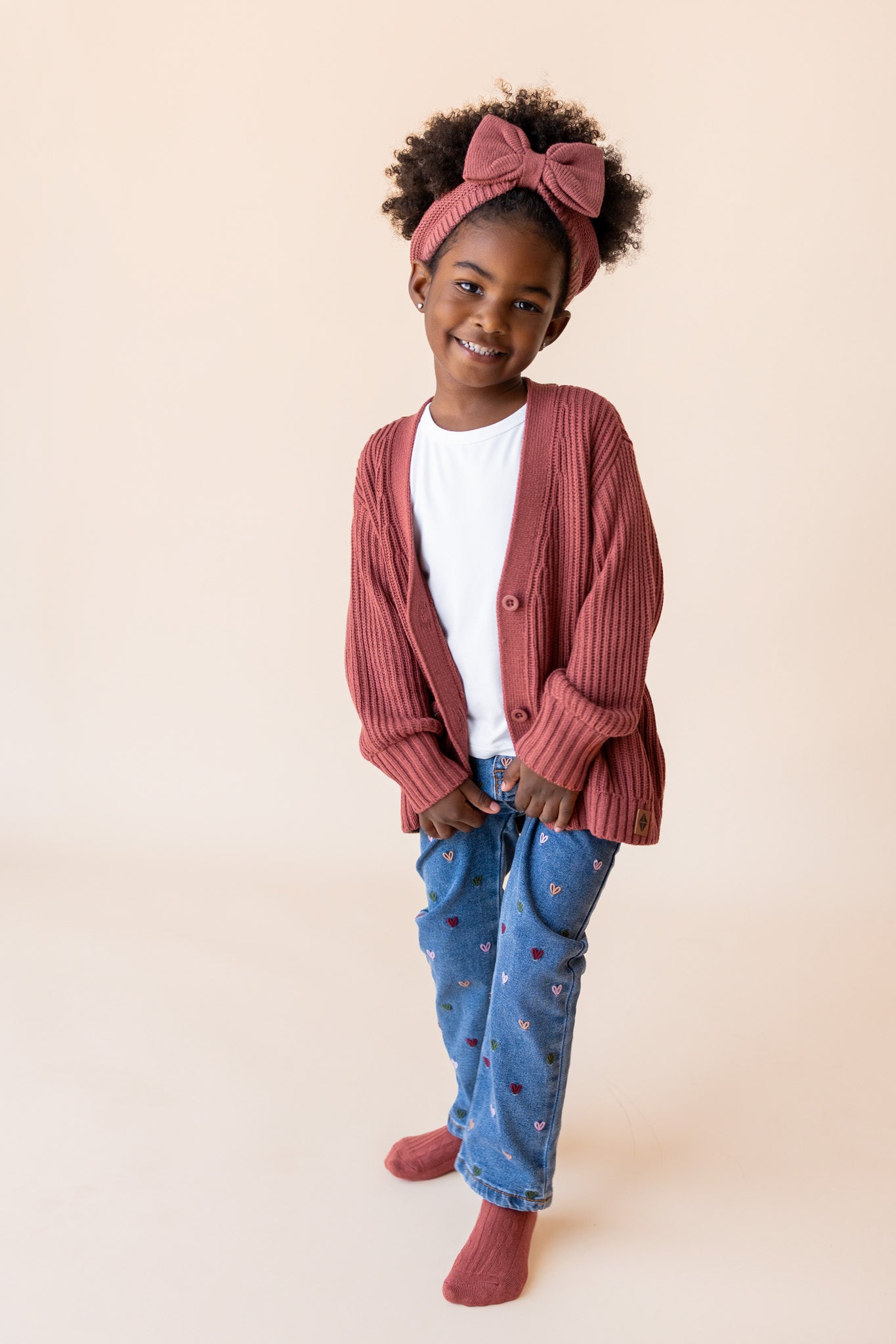 Girl wearing Chunky Knit Oversized Cardigan in Ember over White T-shirt and blue jeans on a beige background