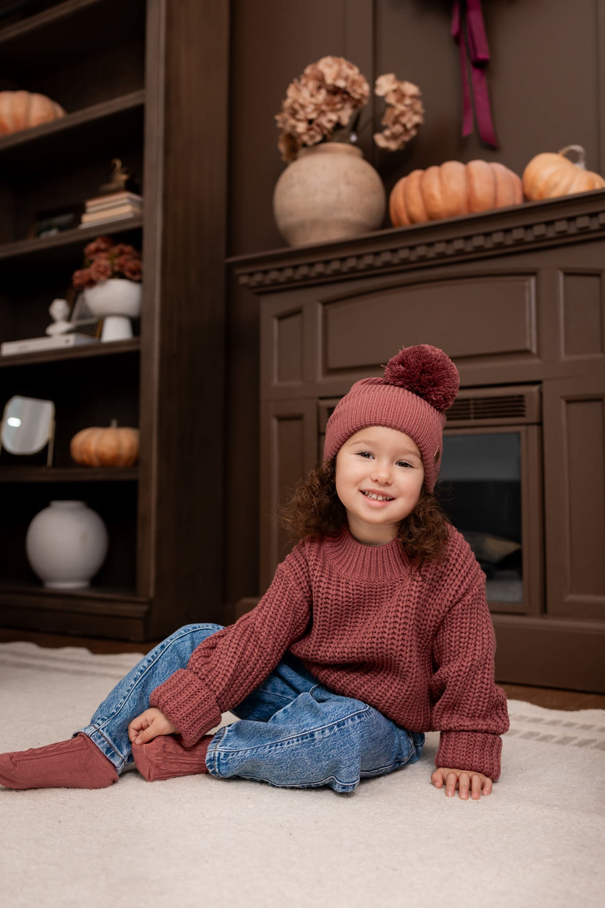 Girl wearing Chunky Knit pom Beanie and chunky knit sweater in Ember over blue jeans sitting on a beige carpet in a living room