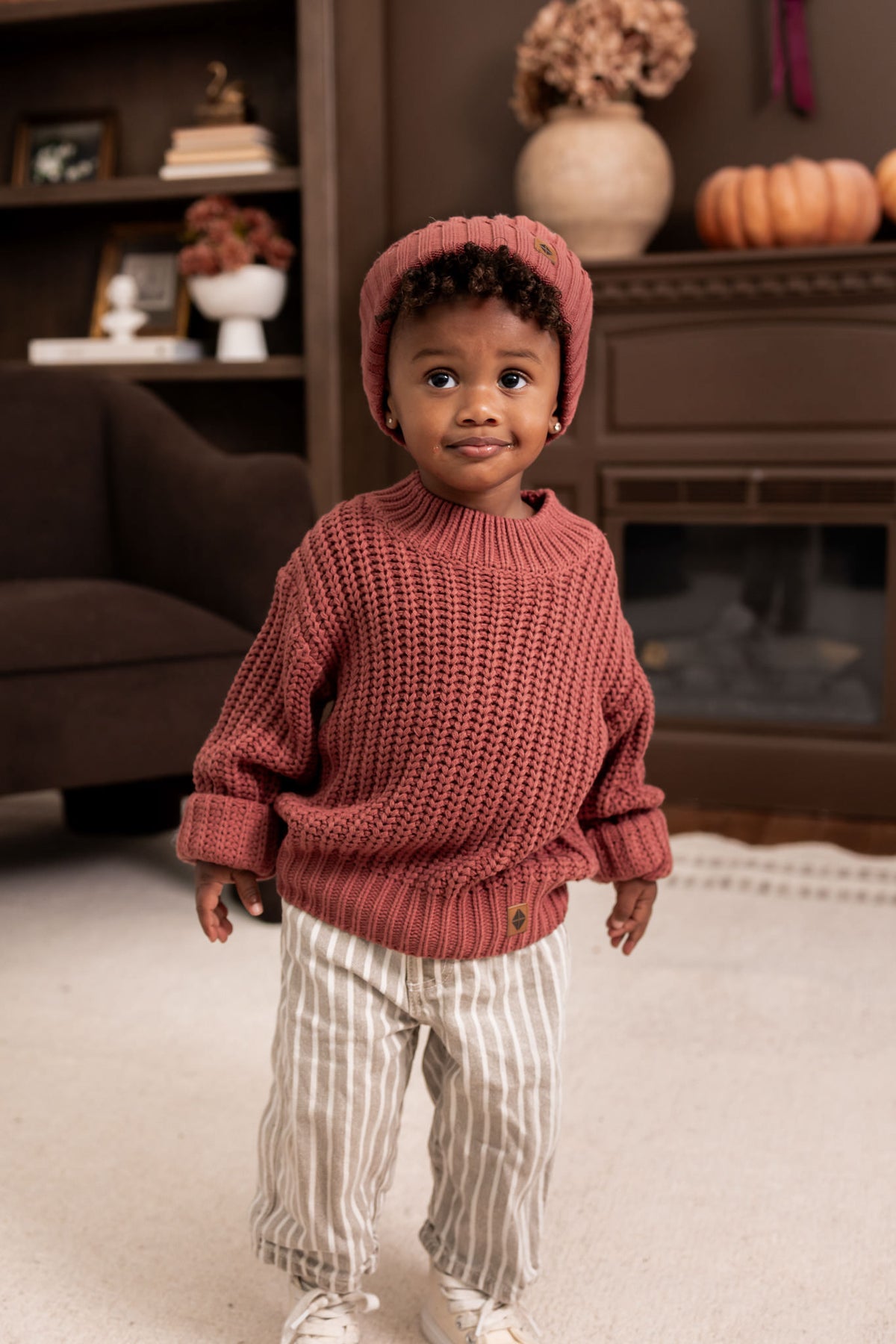 Child standing in a living room wearing Chunky Knit Sweater and chunky knit ribbed beanie in Ember over beige pants