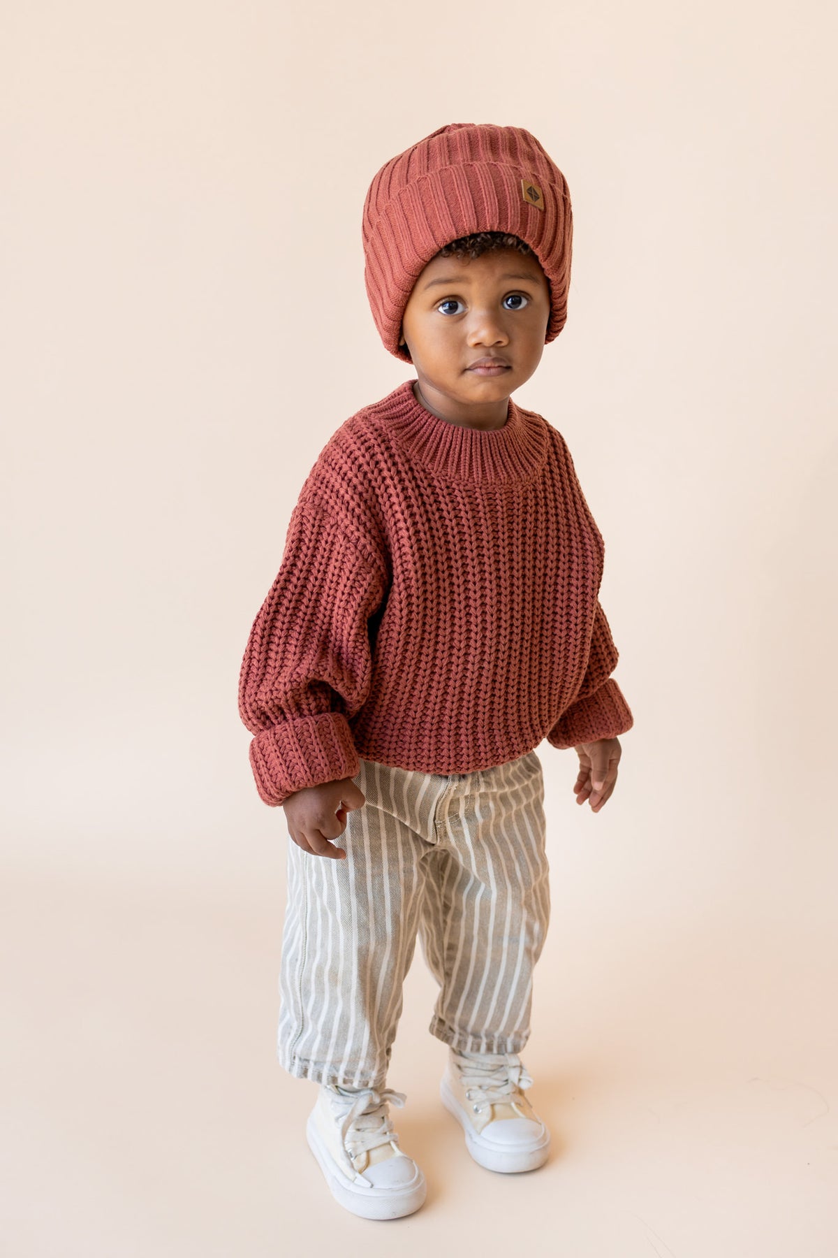 Boy wearing Chunky Knit Sweater and chunky knit ribbed beanie in Ember over beige pants on a beige background