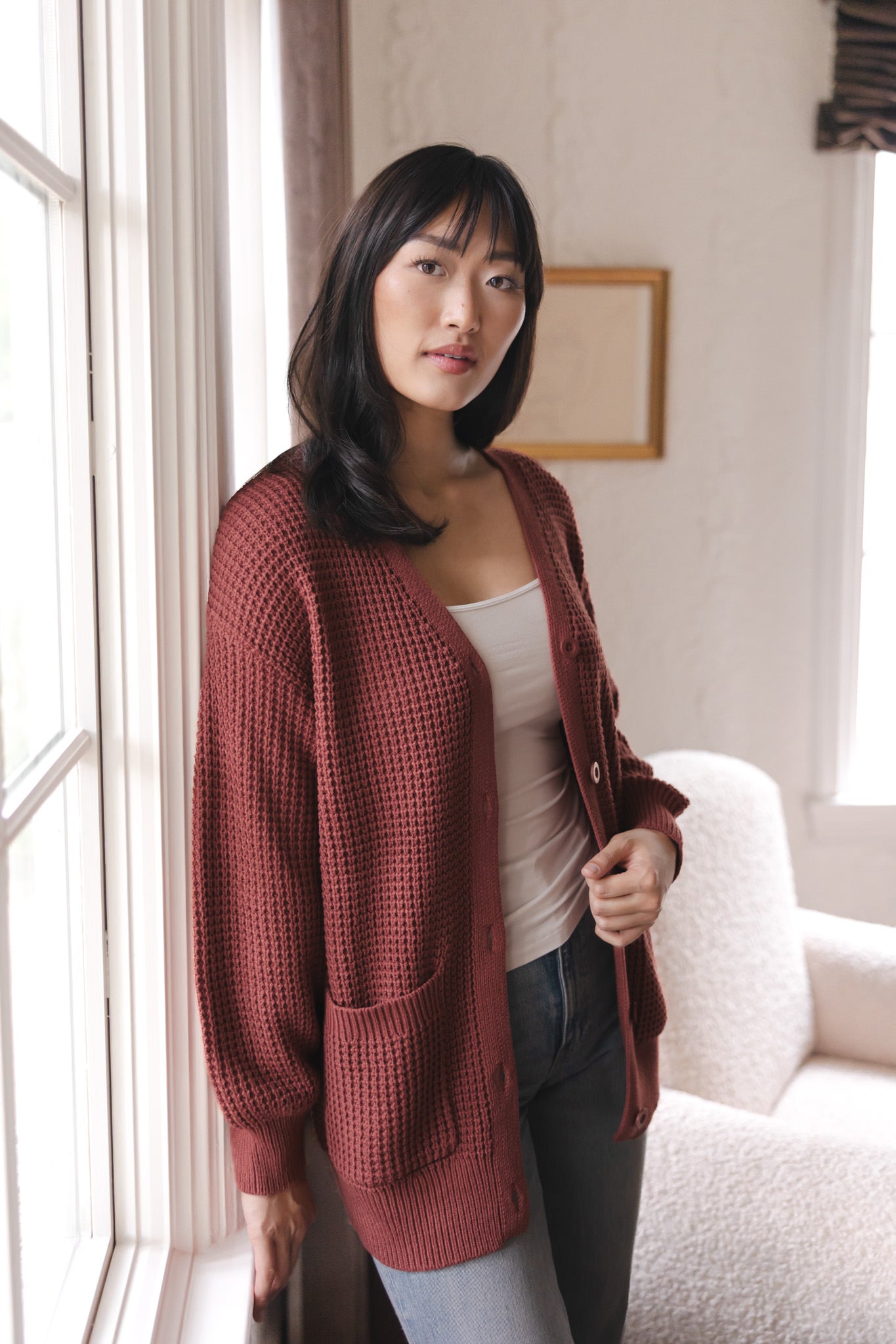Modal posing near a window wearing Chunky Knit Women's Waffle Cardigan in Ember over white camisole and blue jeans