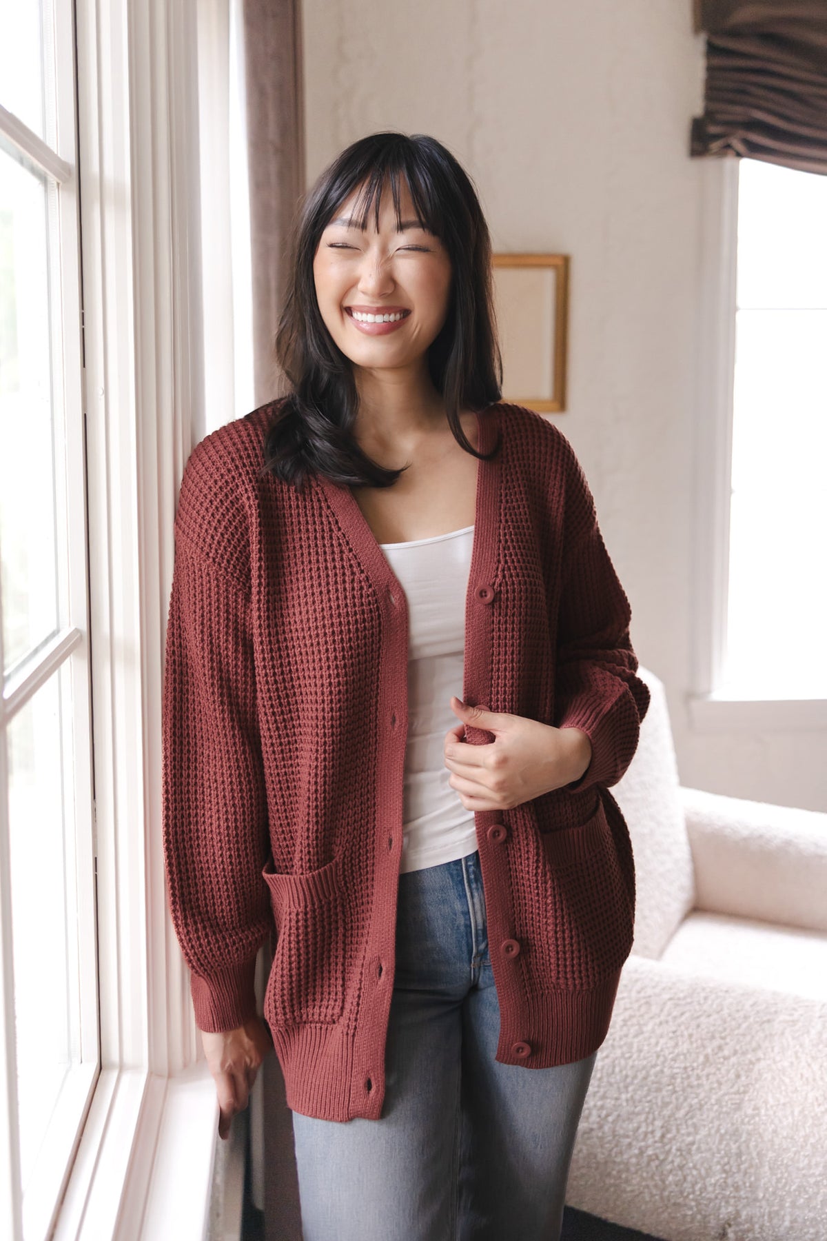 Modal smiling against a window wearing Chunky Knit Women's Waffle Cardigan in Ember over white camisole and blue jeans