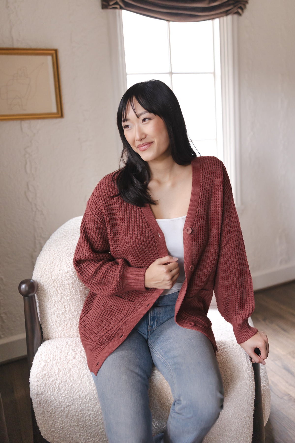 Modal sitting on a white sofa wearing Chunky Knit Women's Waffle Cardigan in Ember over white camisole and blue jeans