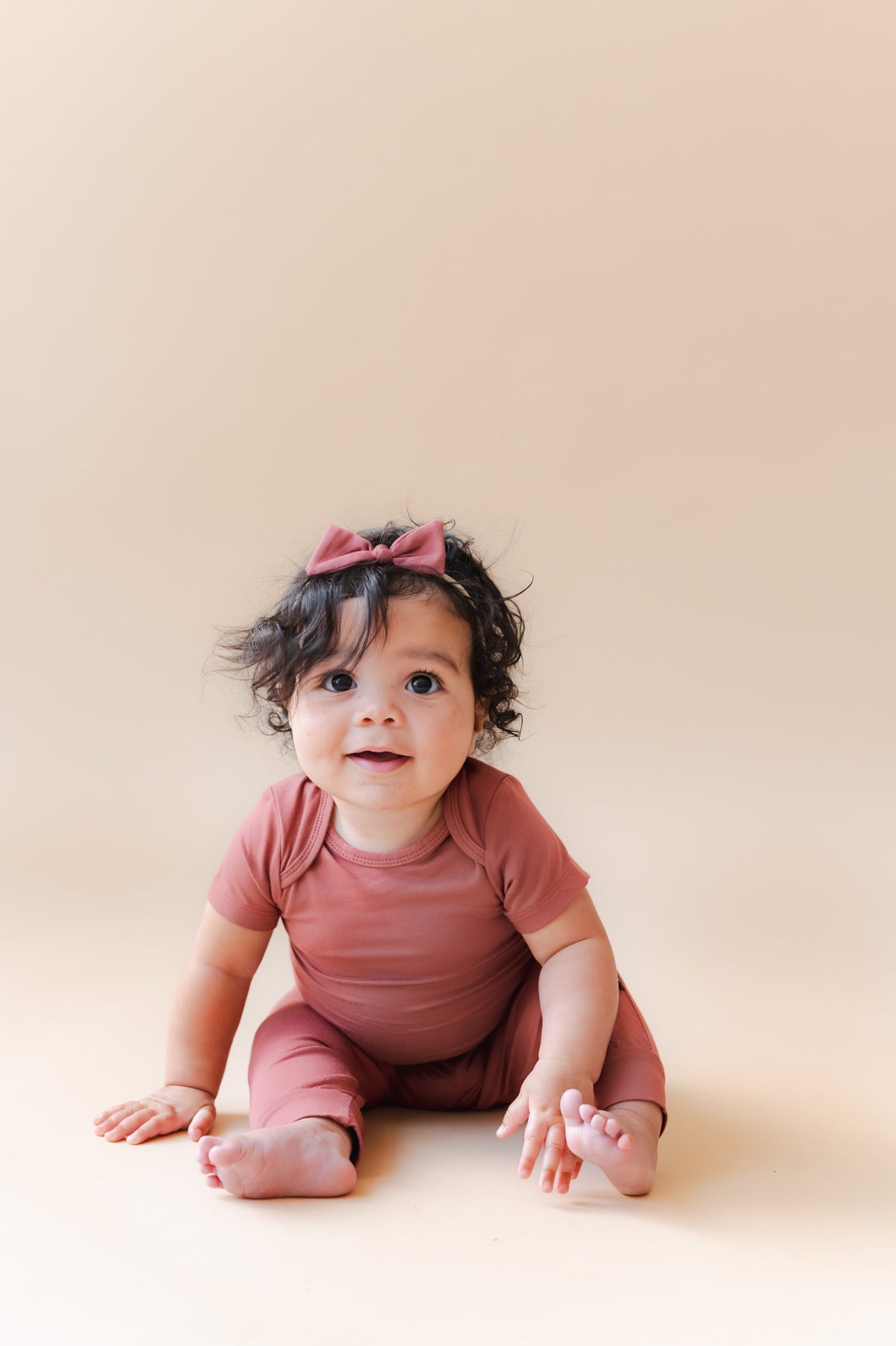 Baby wearing Pant in Ember over similar color bodysuit and bow headband on a beige background