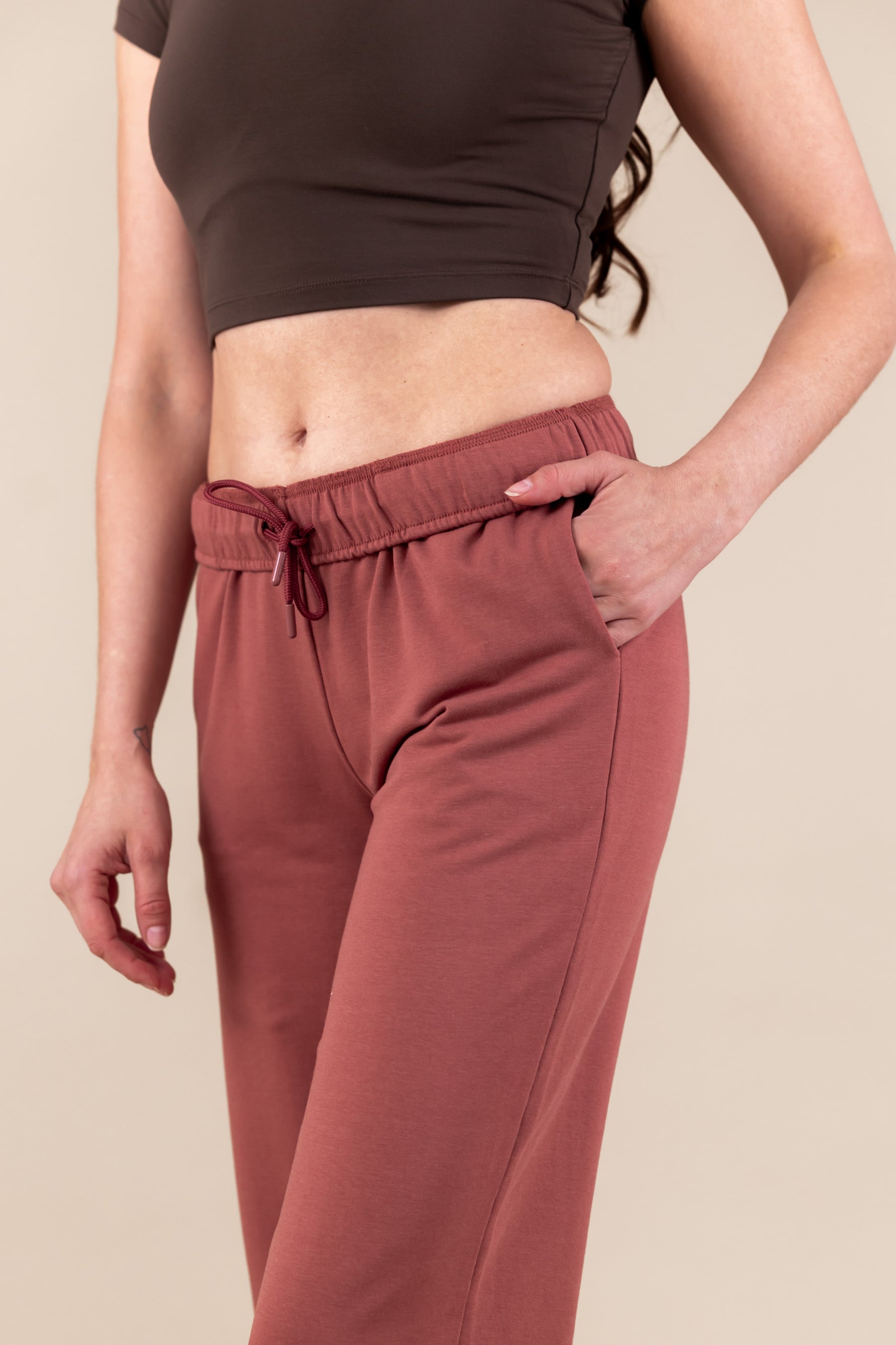 Woman wearing Women's Jogger Pant in Ember color with pockets under a brown shirt on a beige background