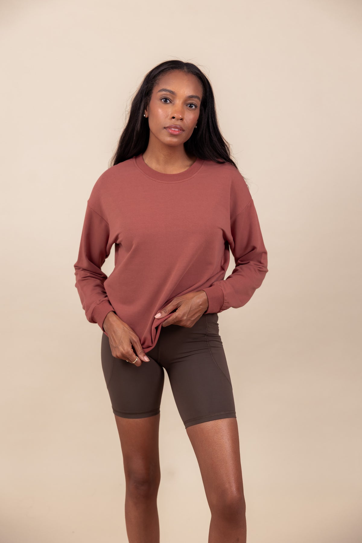 Woman wearing Women's Sweatshirt in Ember color over brown shorts on a beige background