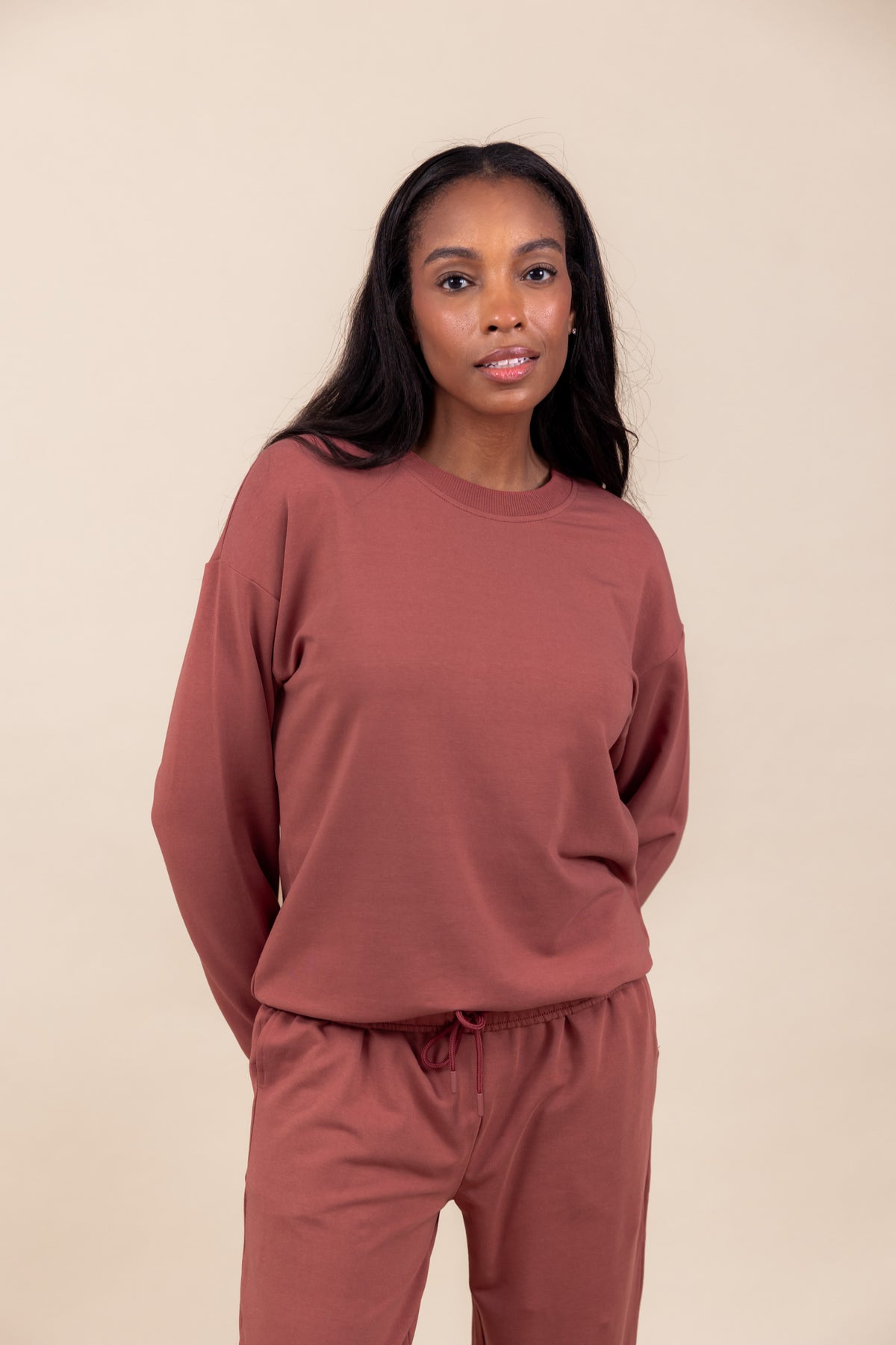 Woman wearing Women's Sweatshirt in Ember color over similar color jogger pants on a beige background