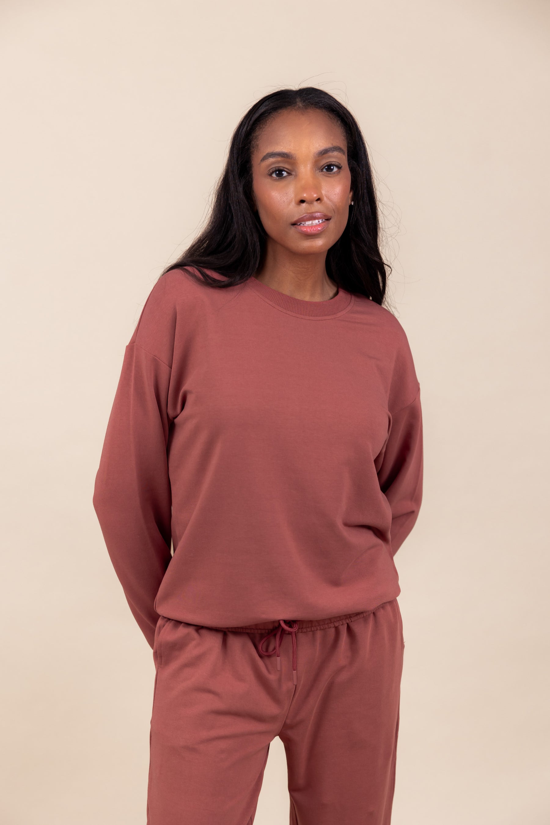 Woman wearing Women's Sweatshirt in Ember color over similar color jogger pants on a beige background