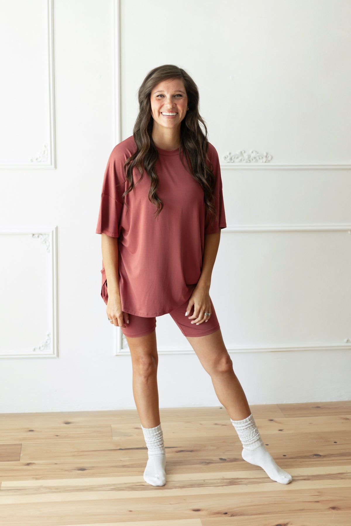 A woman modeling a dusty terracotta  short-sleeve t-shirt and matching shorts set with white socks, standing on a light wood floor against a white paneled wall.