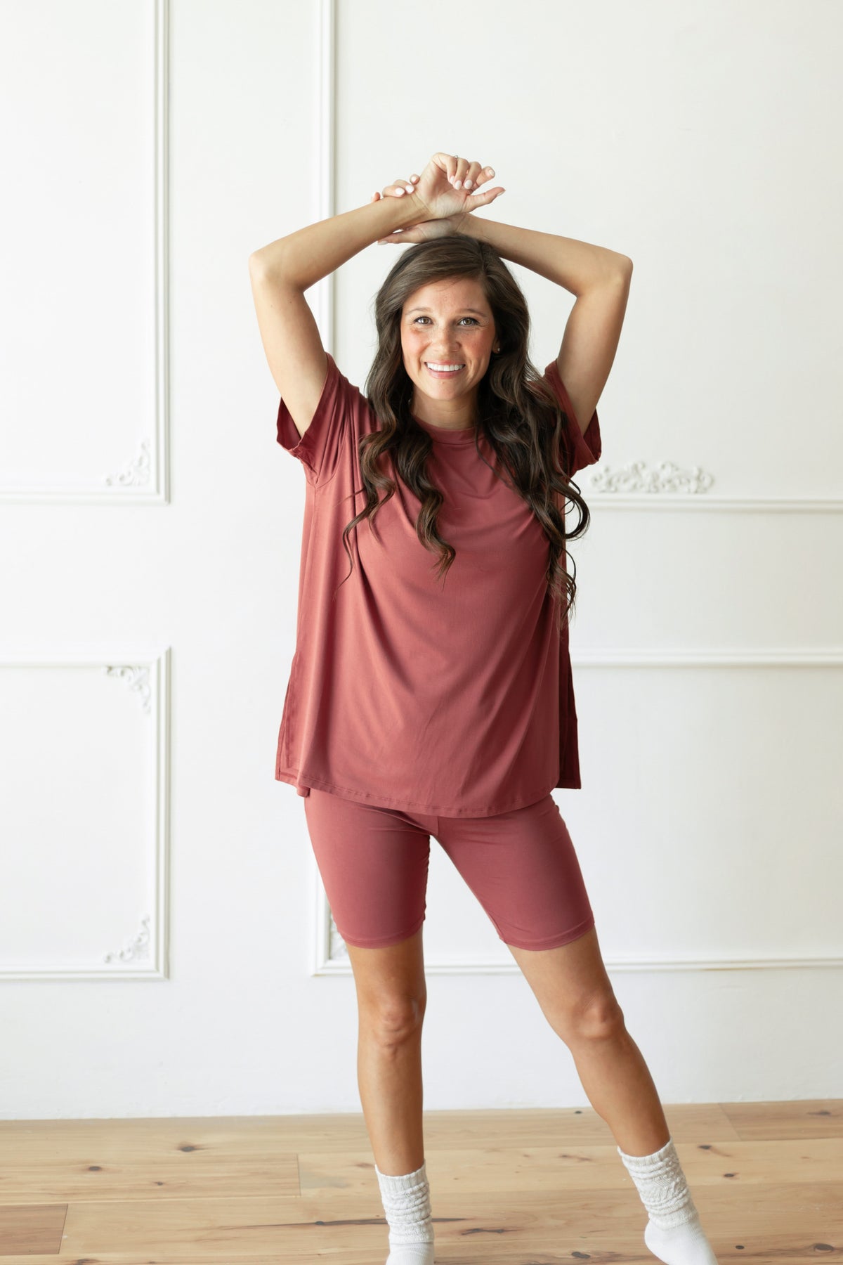 A woman modeling a matching dusty terracotta short-sleeve t-shirt and biker shorts set, standing in a bright room with white paneled walls and light wood floors.