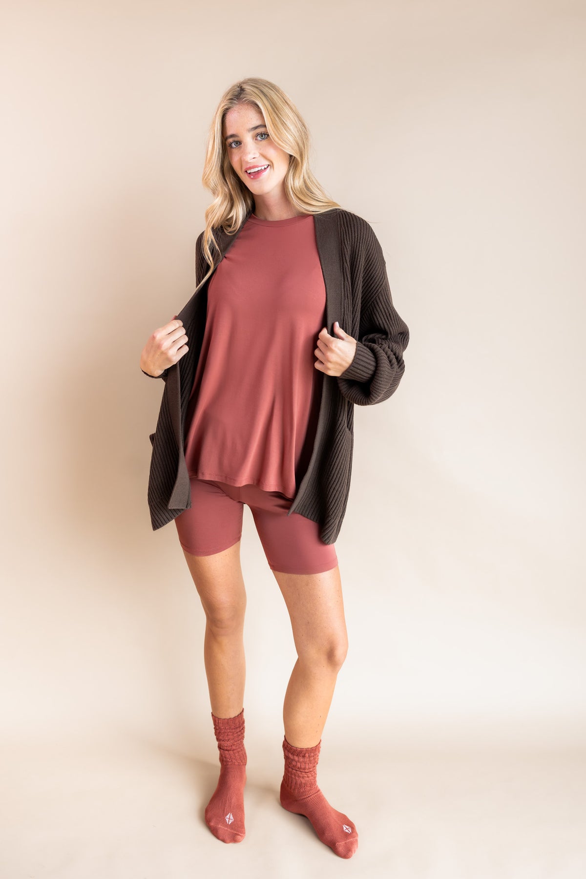 Model wearing Women’s Biker Short Set in Ember with Chunky Knit Oversized Cardigan in Espresso and Adult Socks in Ember on a beige background