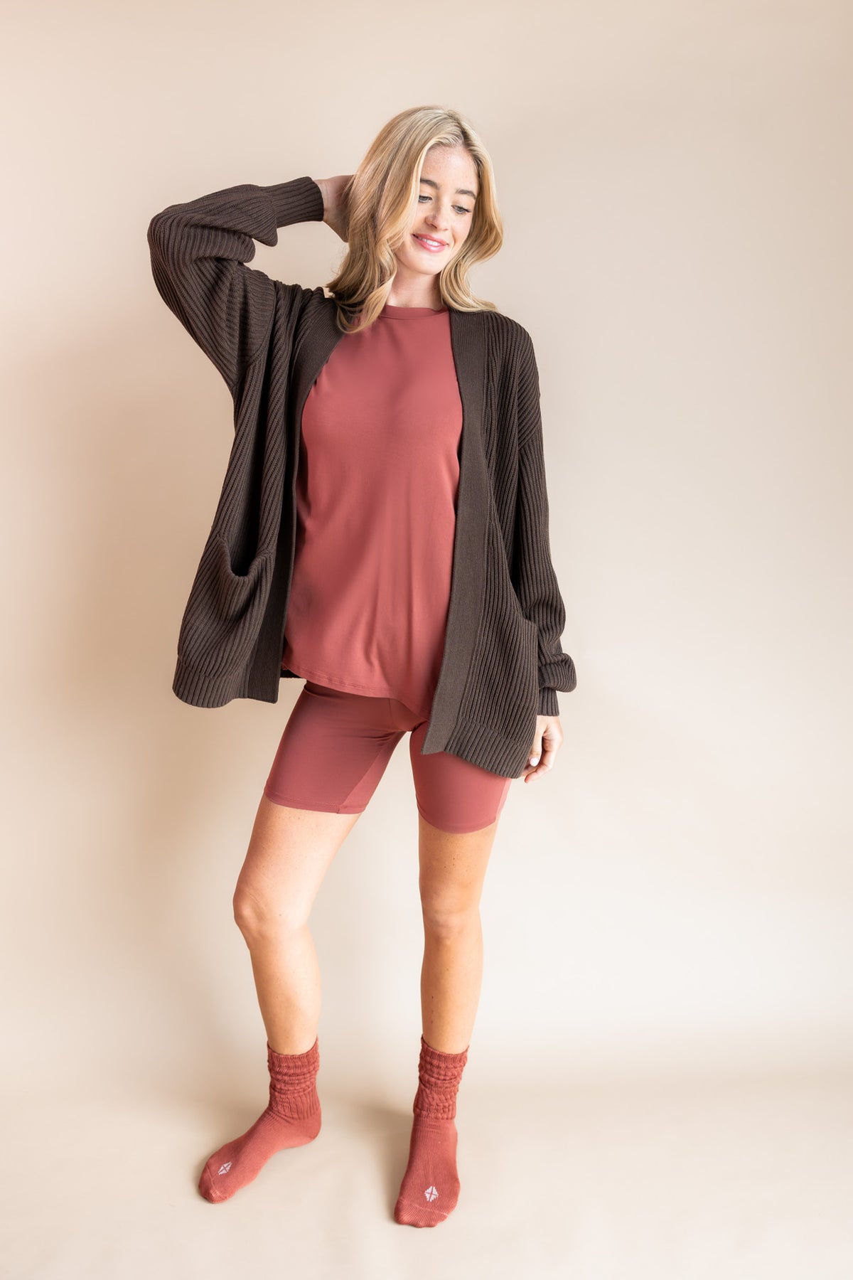 Model posing in Women’s Biker Short Set in Ember with Chunky Knit Oversized Cardigan in Espresso and Adult Socks in Ember on a beige background