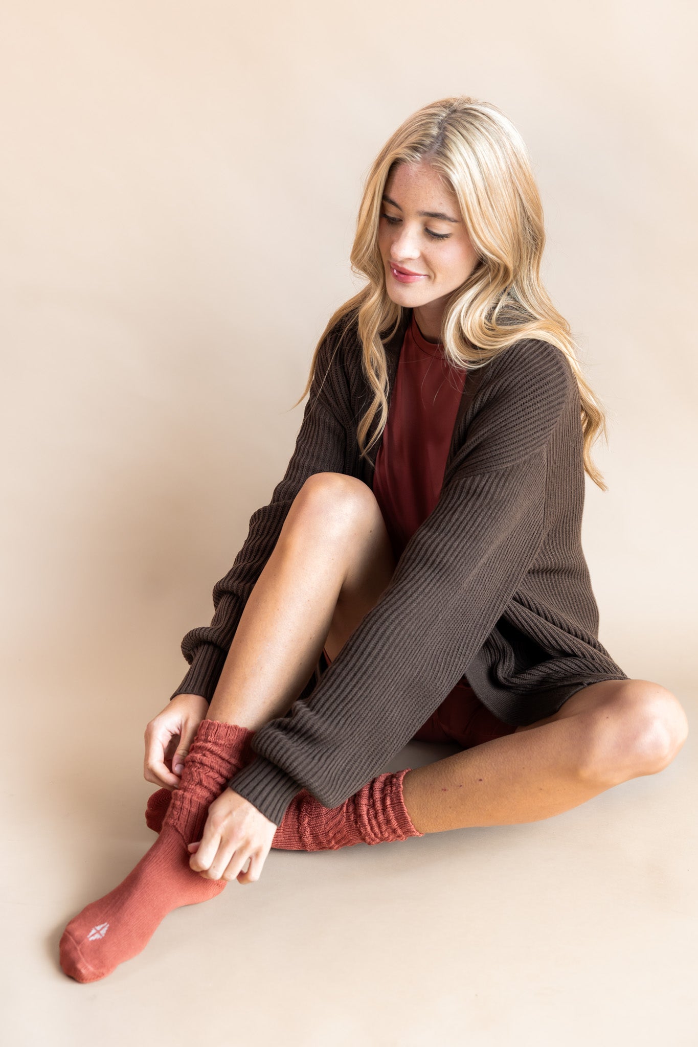 Model sitting while wearing Women’s Biker Short Set in Ember with Chunky Knit Oversized Cardigan in Espresso and Adult Socks in Ember on a beige background