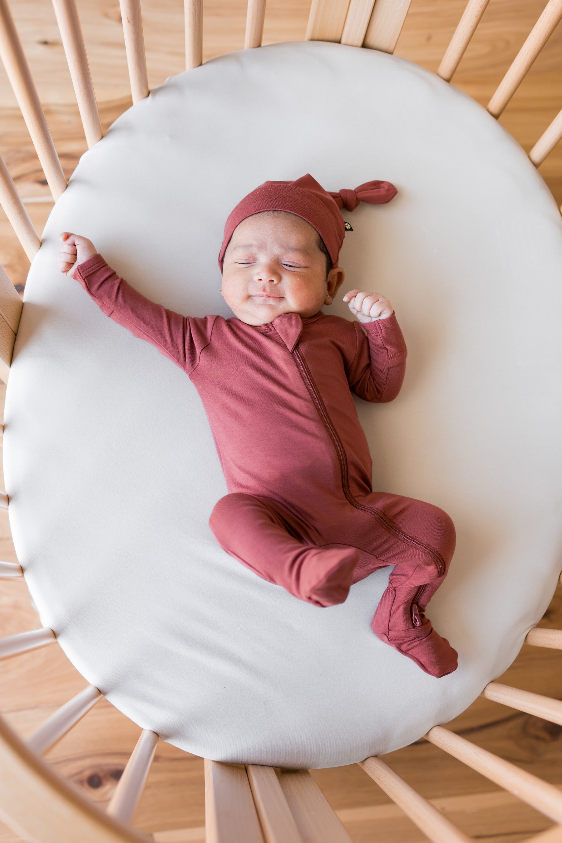 Baby wearing Knotted Cap in Ember with zippered footie in ember lying in a crib