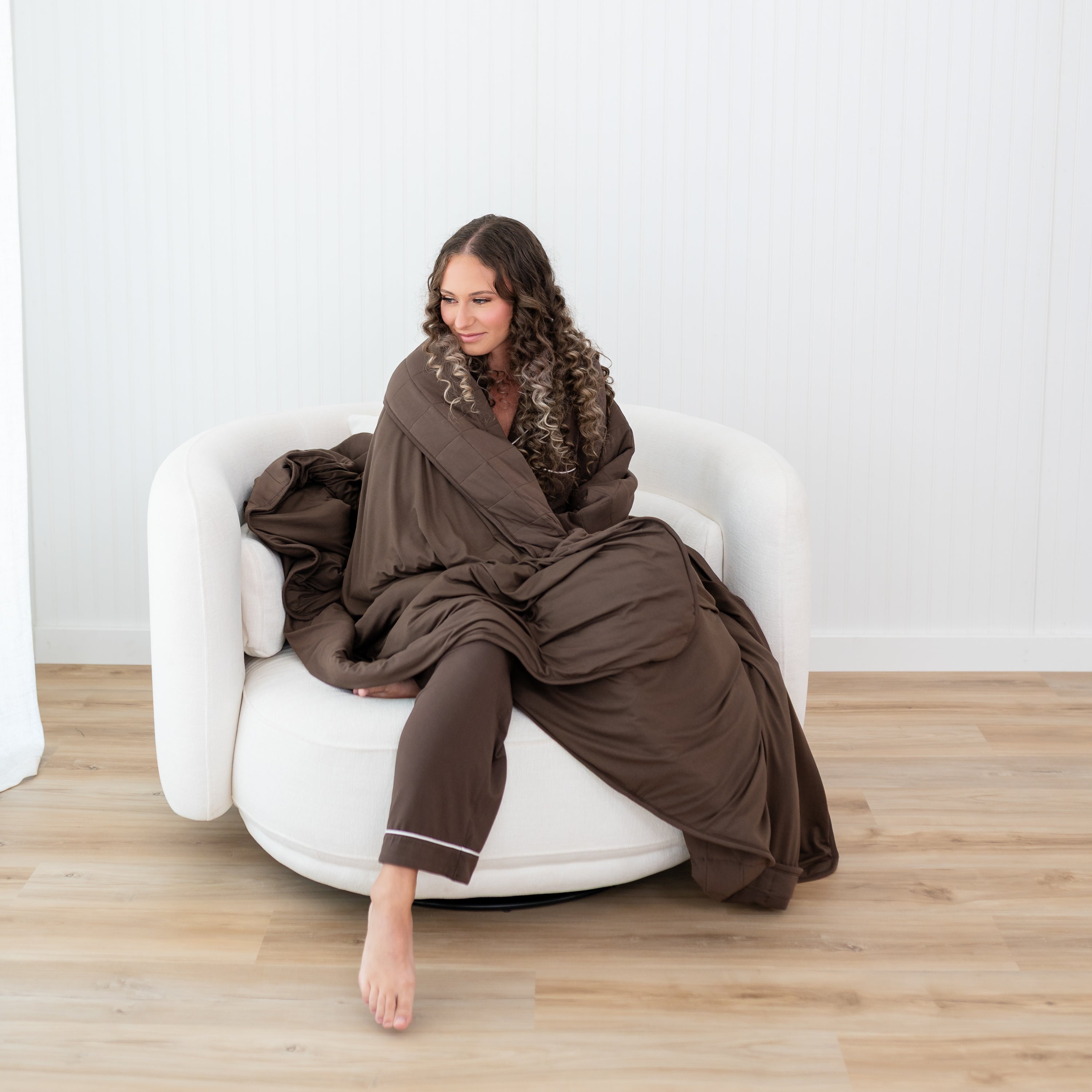Female model sitting on a couch with the Adult Quilted Blanket in Espresso 2.5 wrapped around her