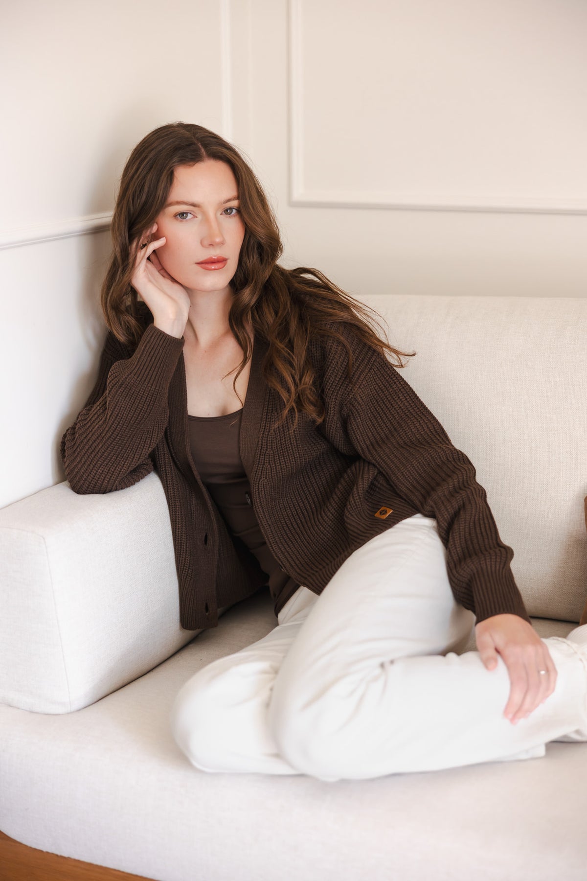 Modal sitting on sofa posing in Chunky Knit Women's Cropped Cardigan in Espresso over espresso camisole and white pants