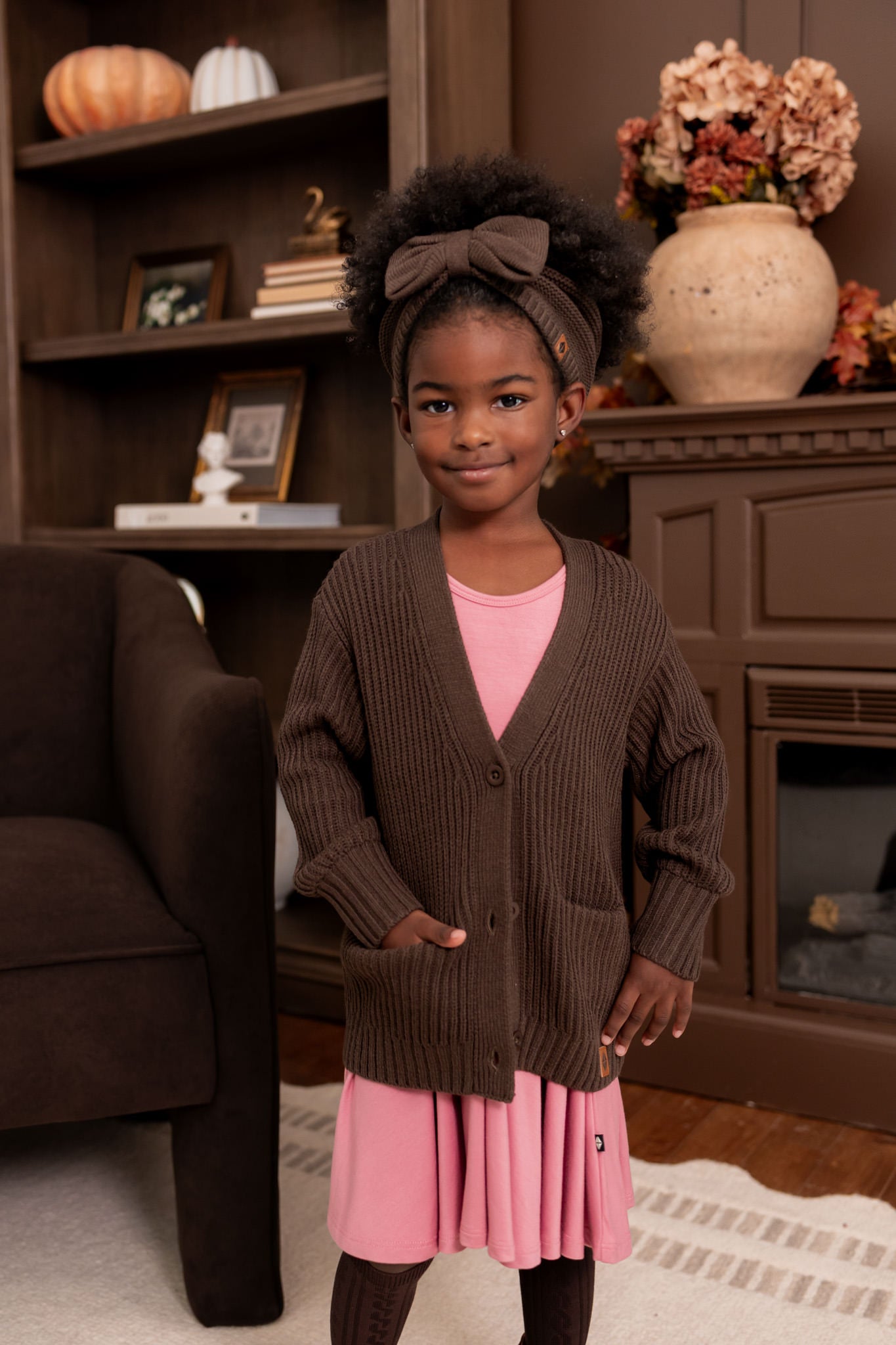Girl wearing Chunky Knit Oversized Cardigan in Espresso over Twirl dress in pink and chunky knit bow headband in espresso standing in a living room