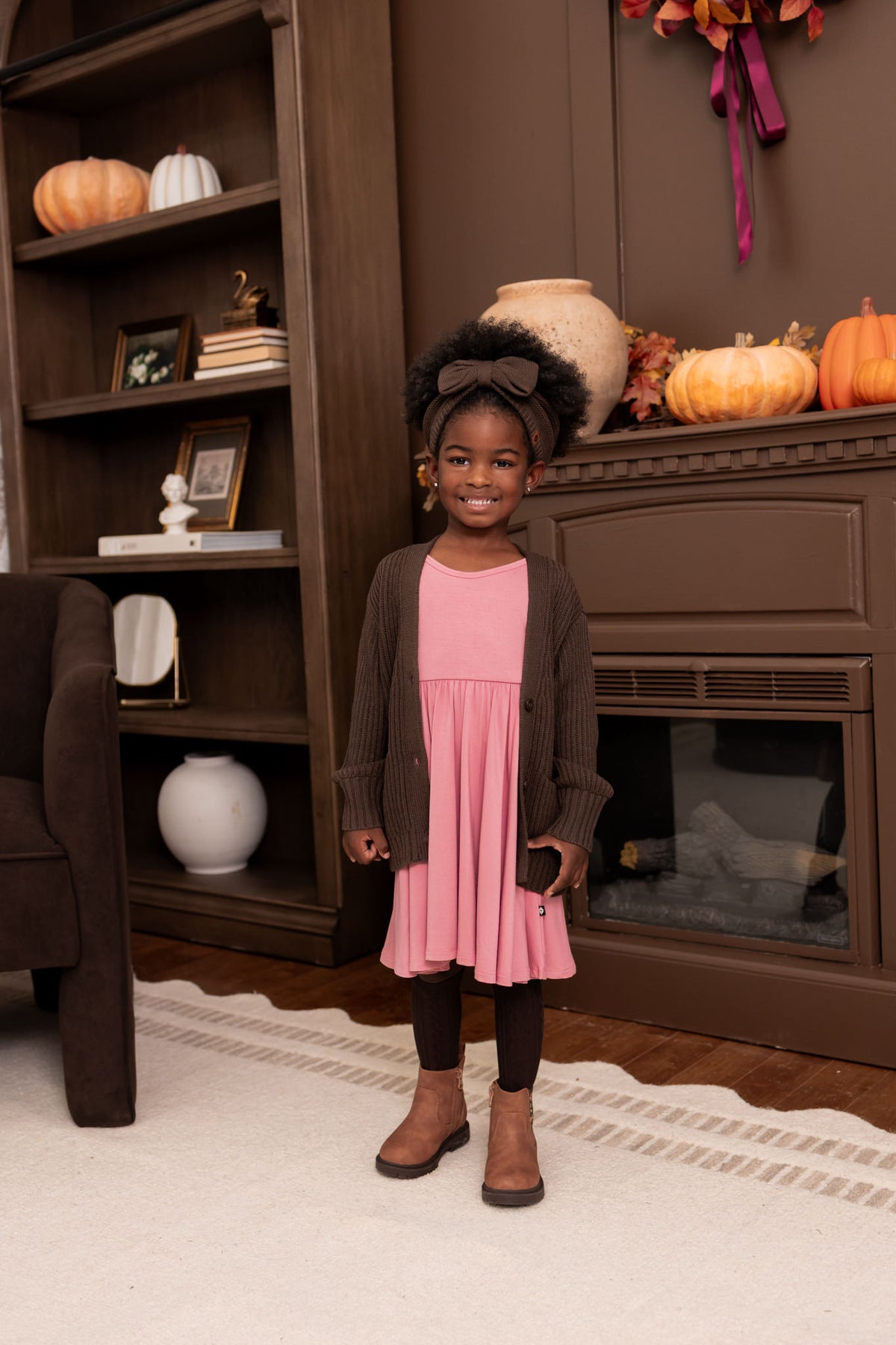 Girl wearing Chunky Knit Oversized Cardigan in Espresso over Twirl dress in pink  and chunky knit bow headband in espresso standing in a living room