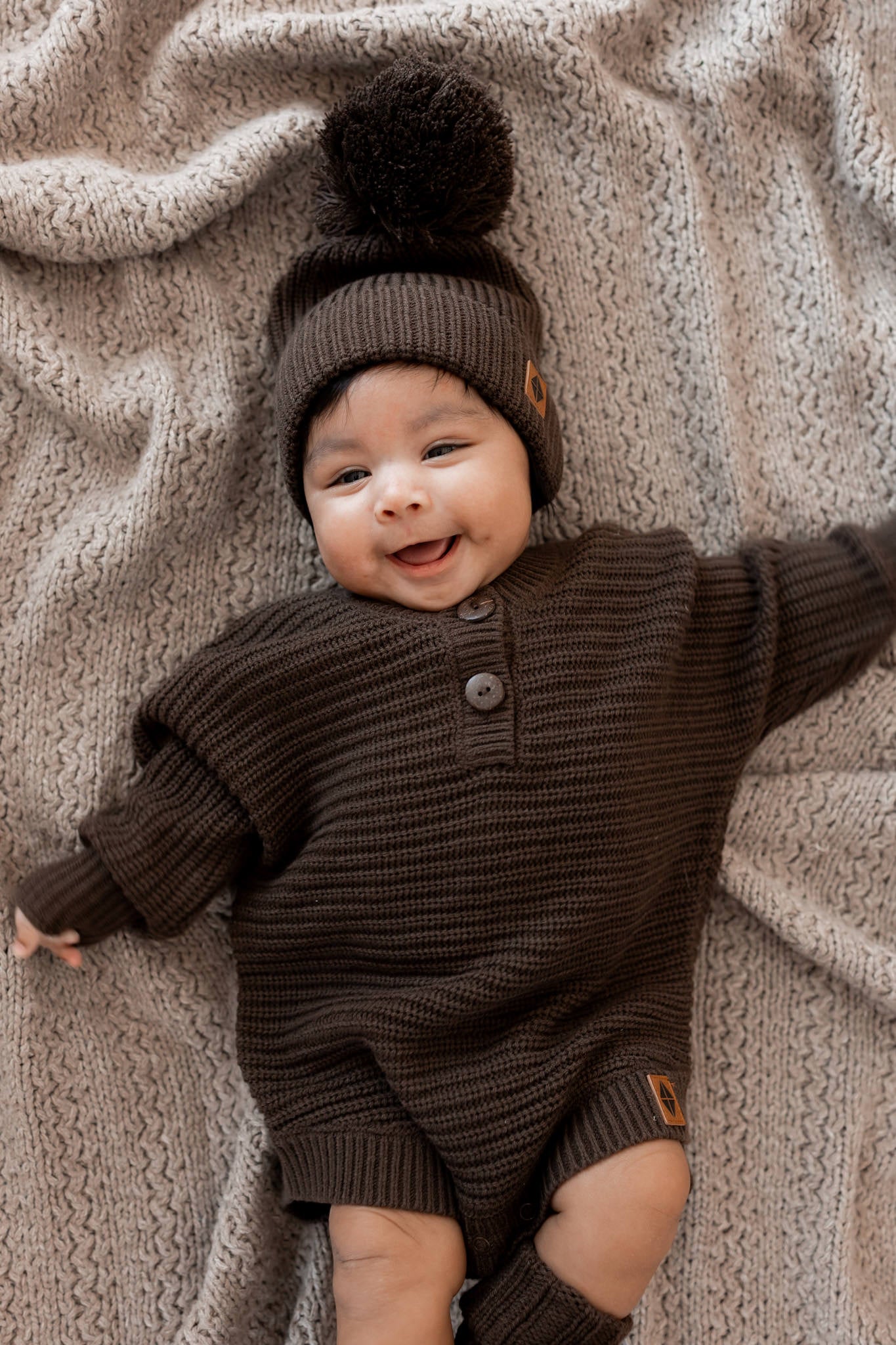 Baby smiling while lying on a beige blanket wearing Chunky Knit Pom Beanie, chunky knit romper and knit booties in Espresso