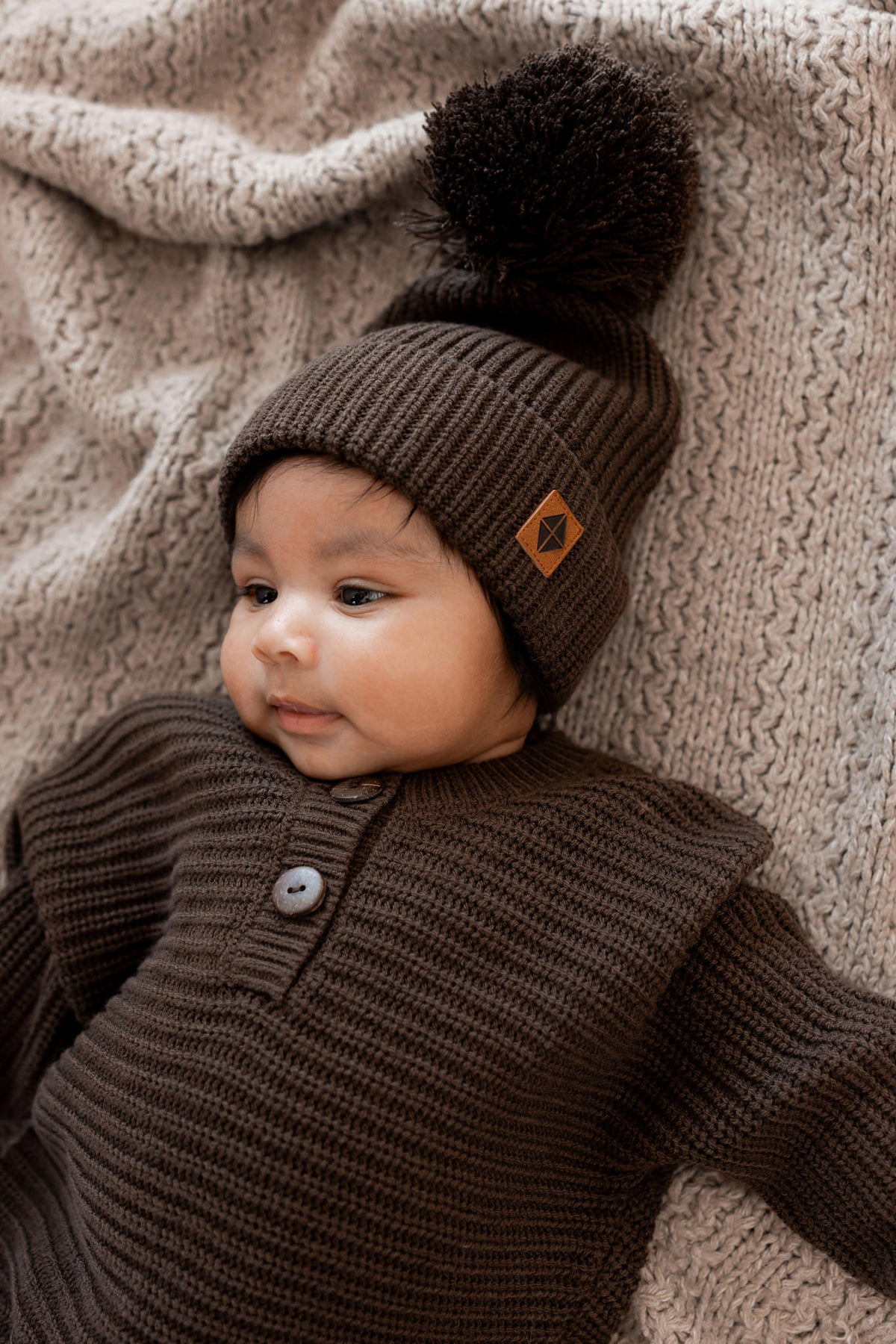 Baby wearing Chunky Knit Romper and chunky knit pom beanie in Espresso on a beige blanket