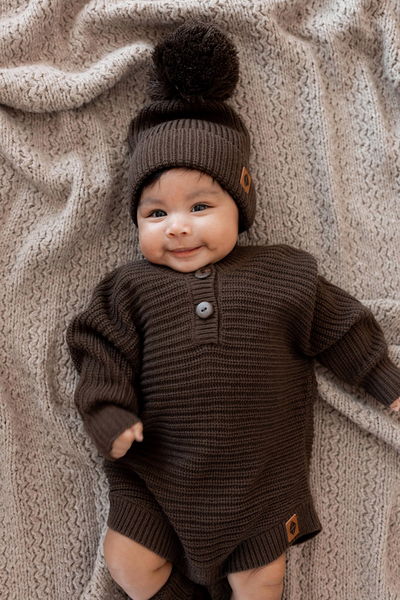 Baby smiling in Chunky Knit Romper and chunky knit pom beanie in Espresso lying on a beige blanket
