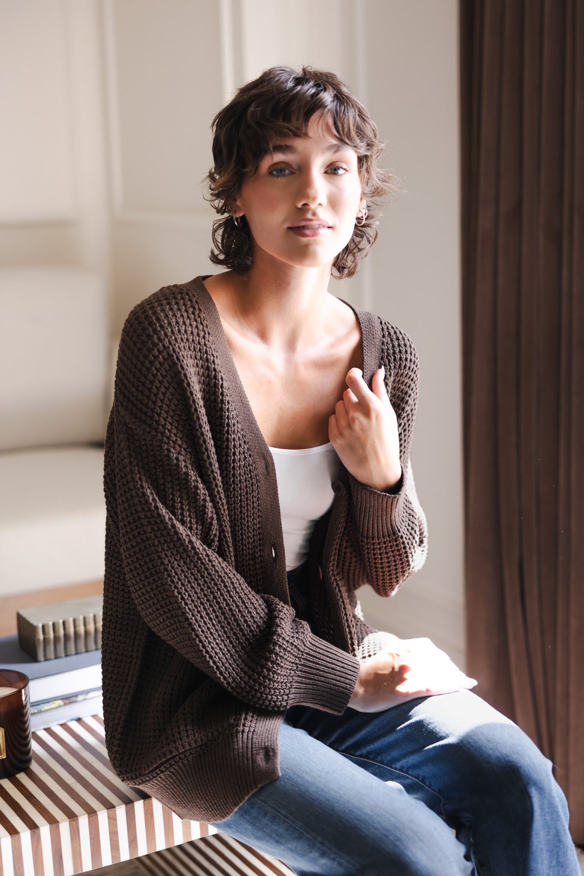 Modal sitting on a table wearing Chunky Knit Women's Waffle Cardigan in Espresso over white camisole and blue jeans