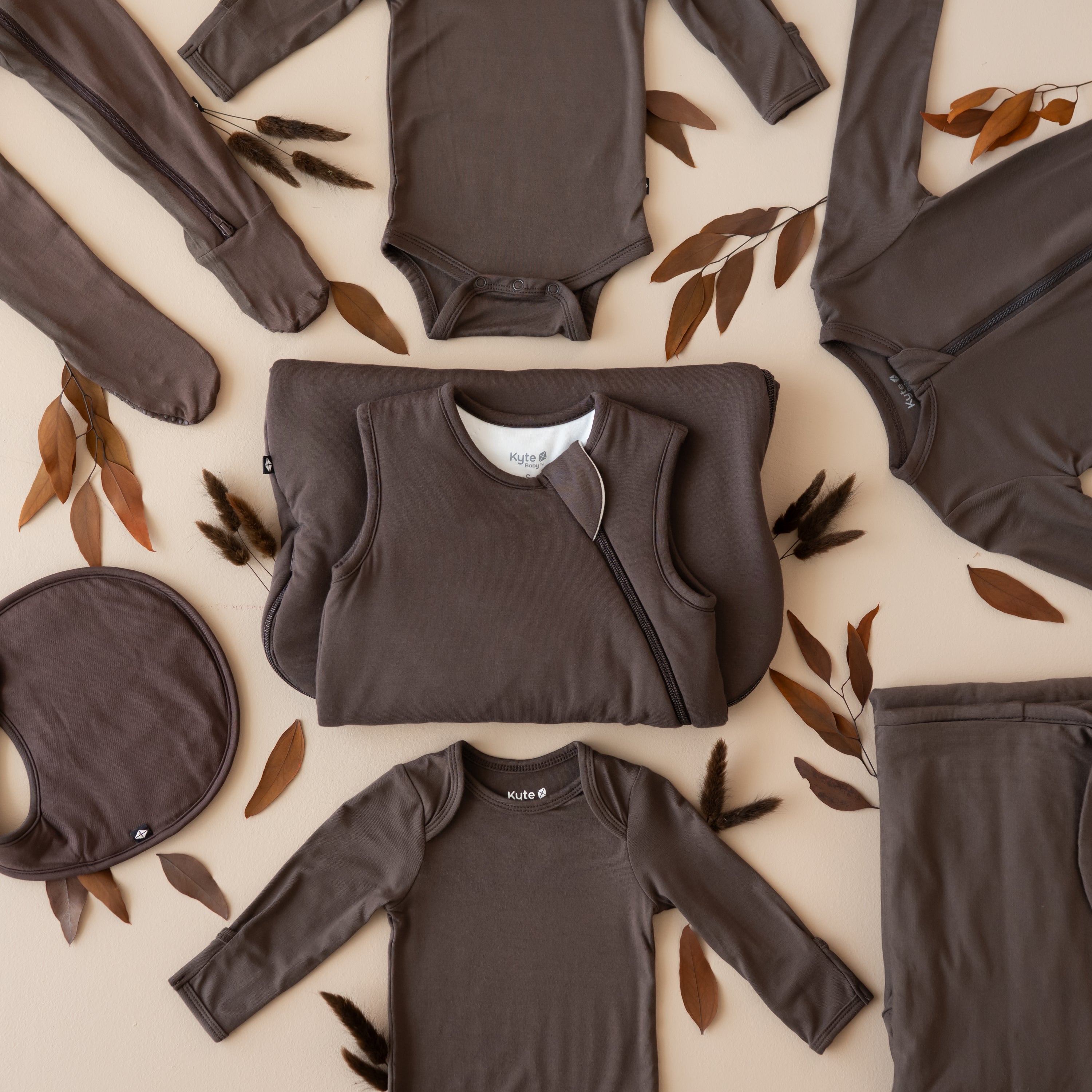 Espresso flat lay with the Sleep Bag in 1.0 in the middle surrounded by a long sleeve bodysuits, zippered footie, cushy bib and baby blanket
