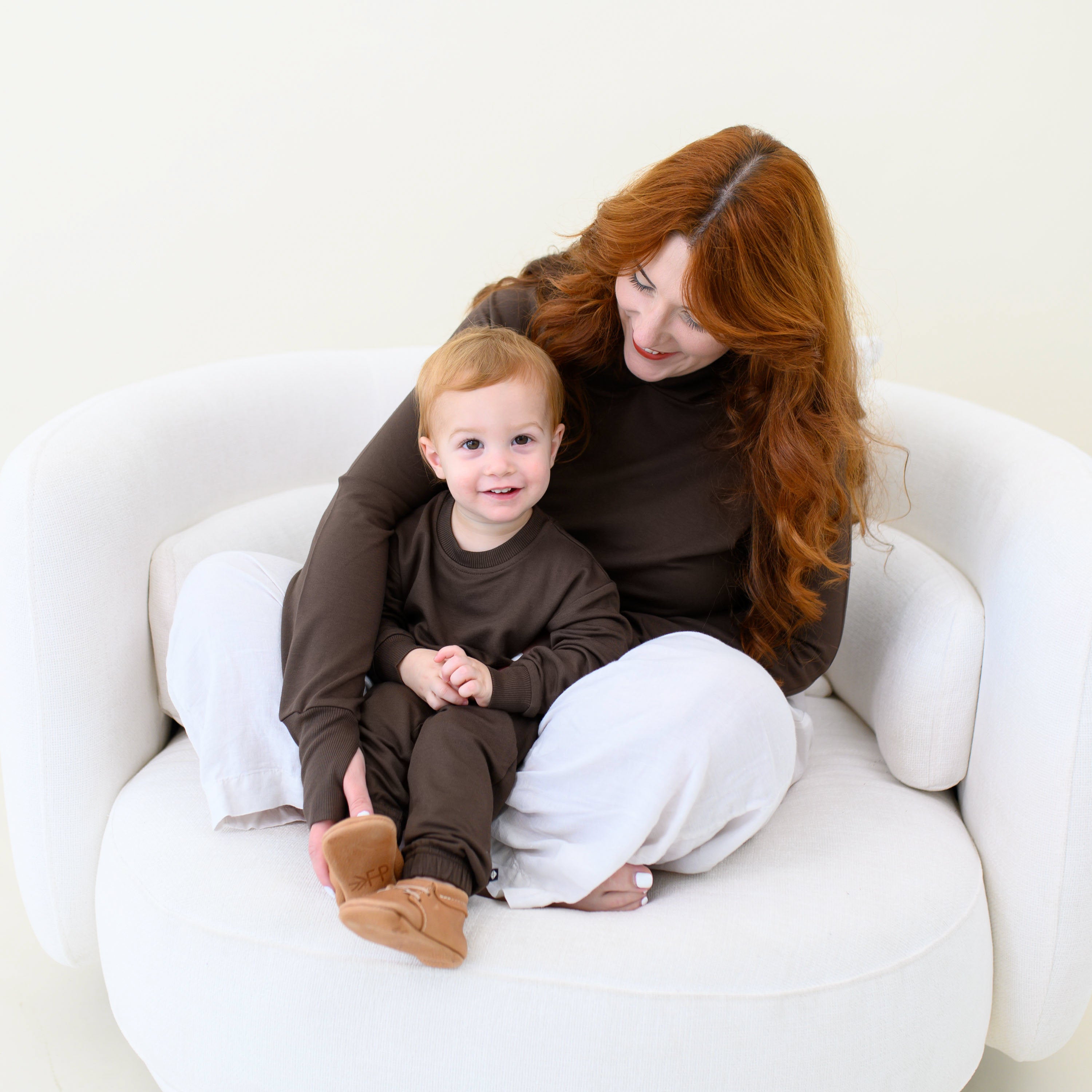 Mother and son sitting on a cream colored sofa matching in Espresso. The boy is wearing the French Terry Jogger Set in Espresso while the mother is wearing the French Terry Nursing Hoodie