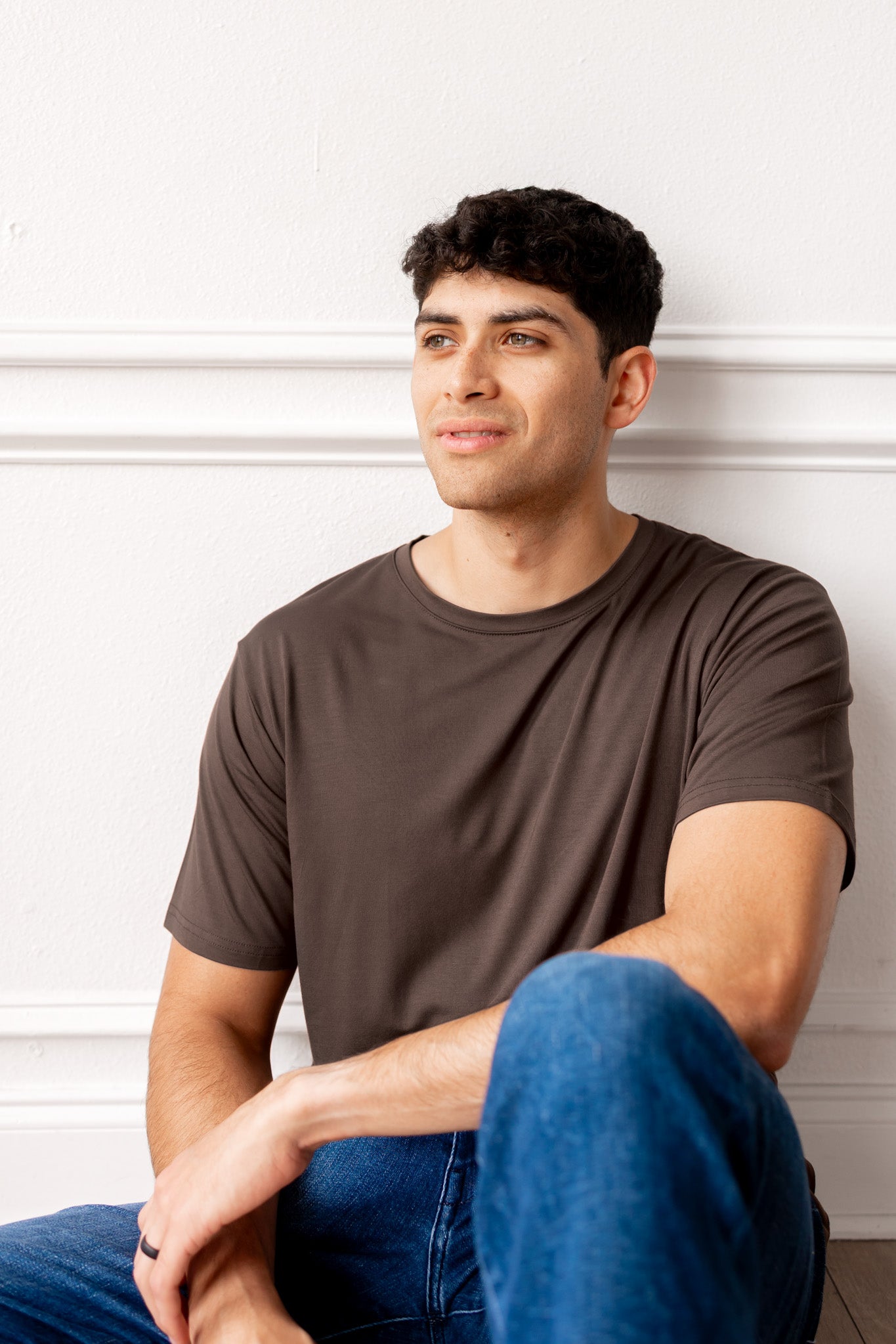 Man sitting on a floor wearing a brown t-shirt and blue jeans a white paneled wall.