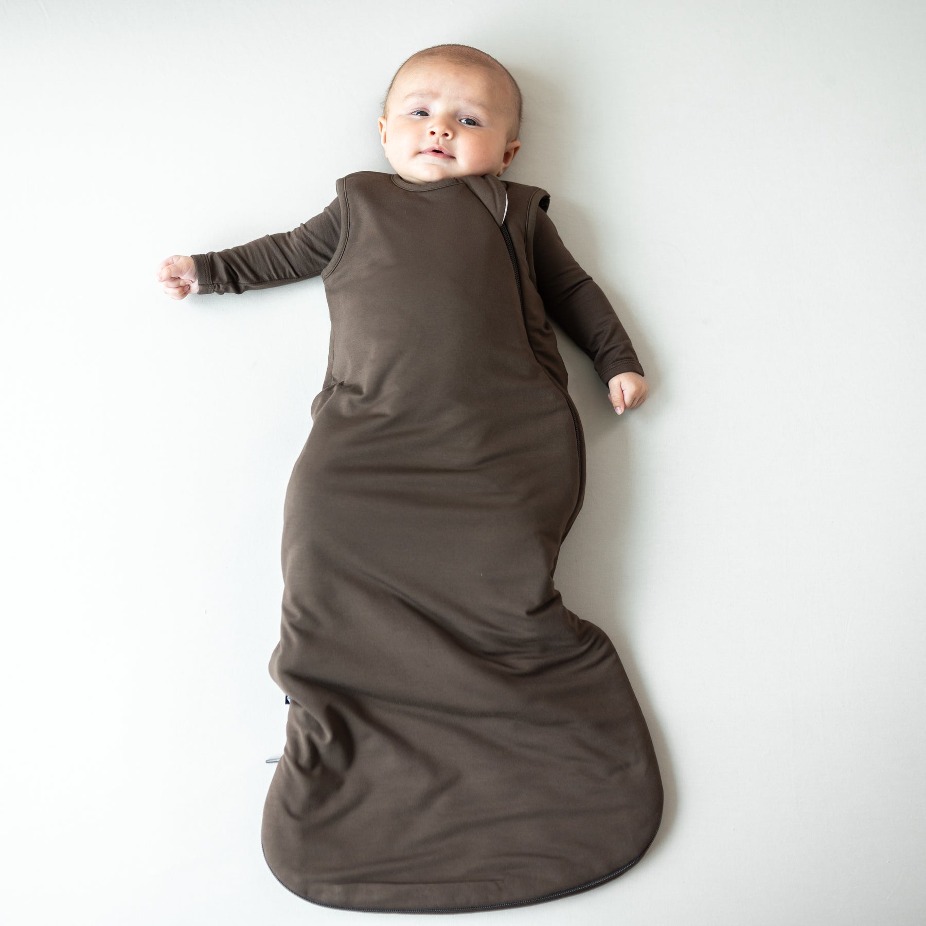 Infant laying down wearing the Sleep Bag in Espresso 1.0 paired with a matching long sleeve romper on underneath