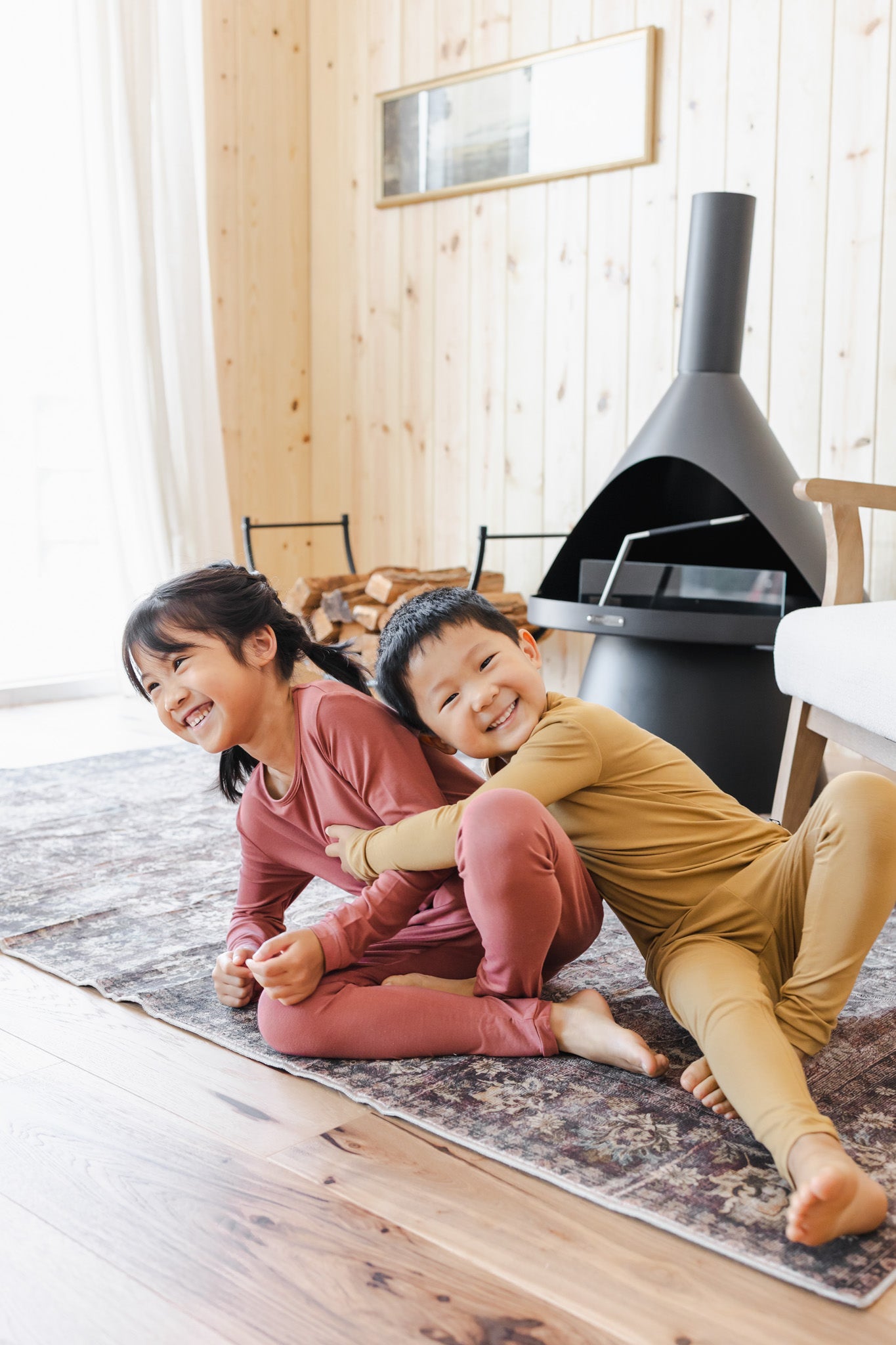 Two children sitting on a patterned rug in a cozy room with wood-paneled walls. The girl wears a dusty rose long-sleeve pajama set, and the boy wears a mustard yellow long-sleeve pajama set.