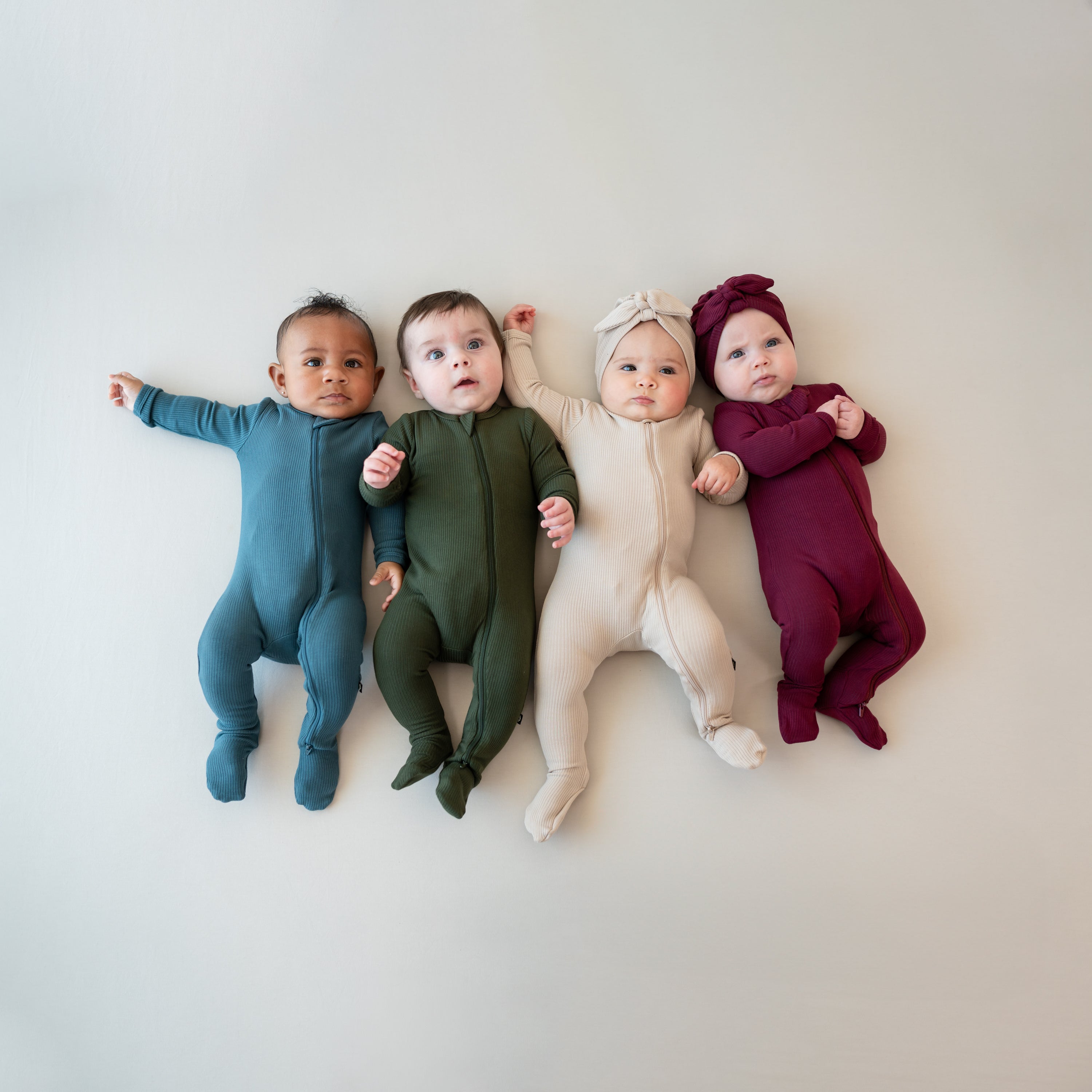 Four infant models laying side by side wearing the Ribbed Zipper Footie in Atlantic, Fir, Bisque and Burgundy