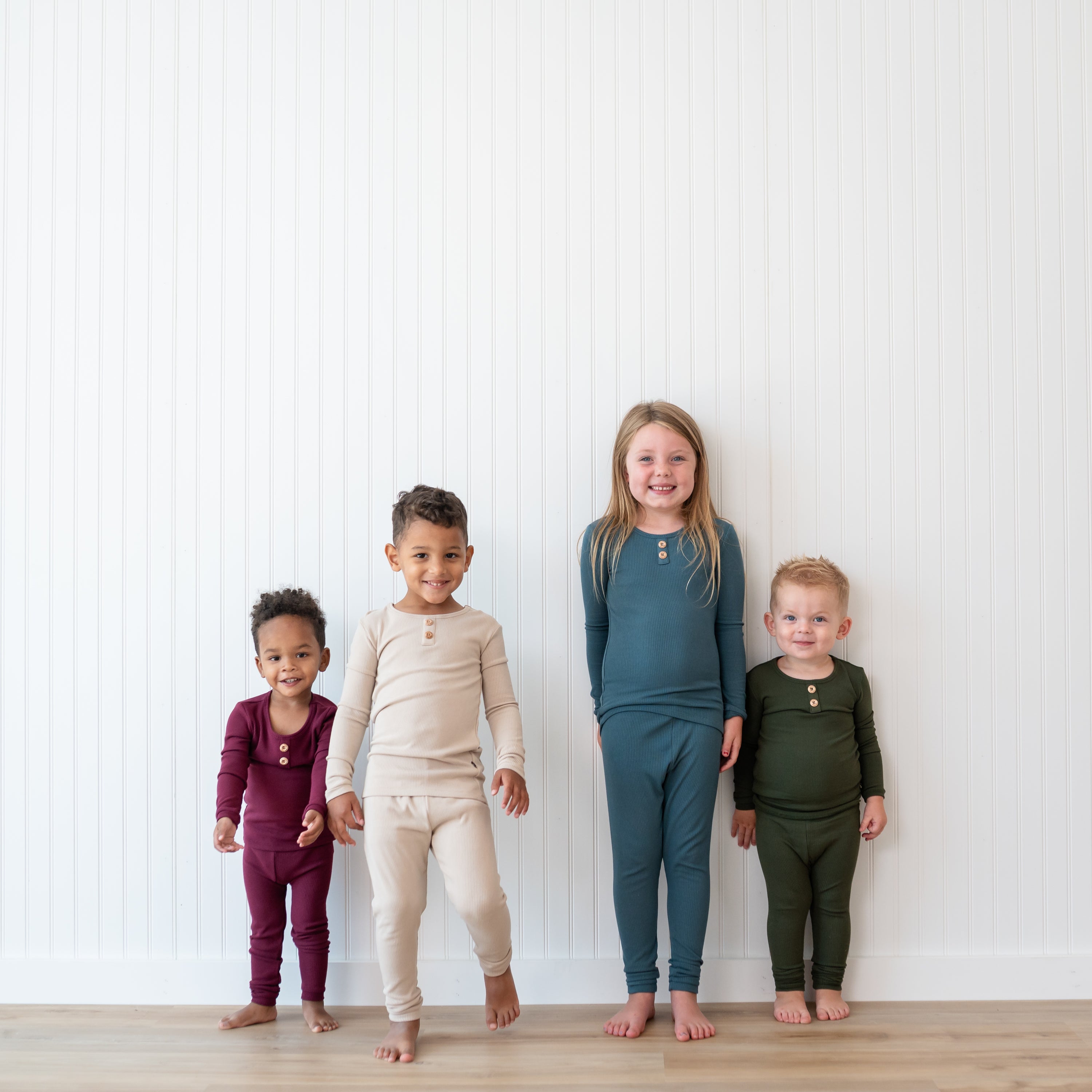 Four children standing side by side in front of a white paneled wall wearing the Ribbed Henley Sets in Burgundy, Bisque, Atlantic and Fir
