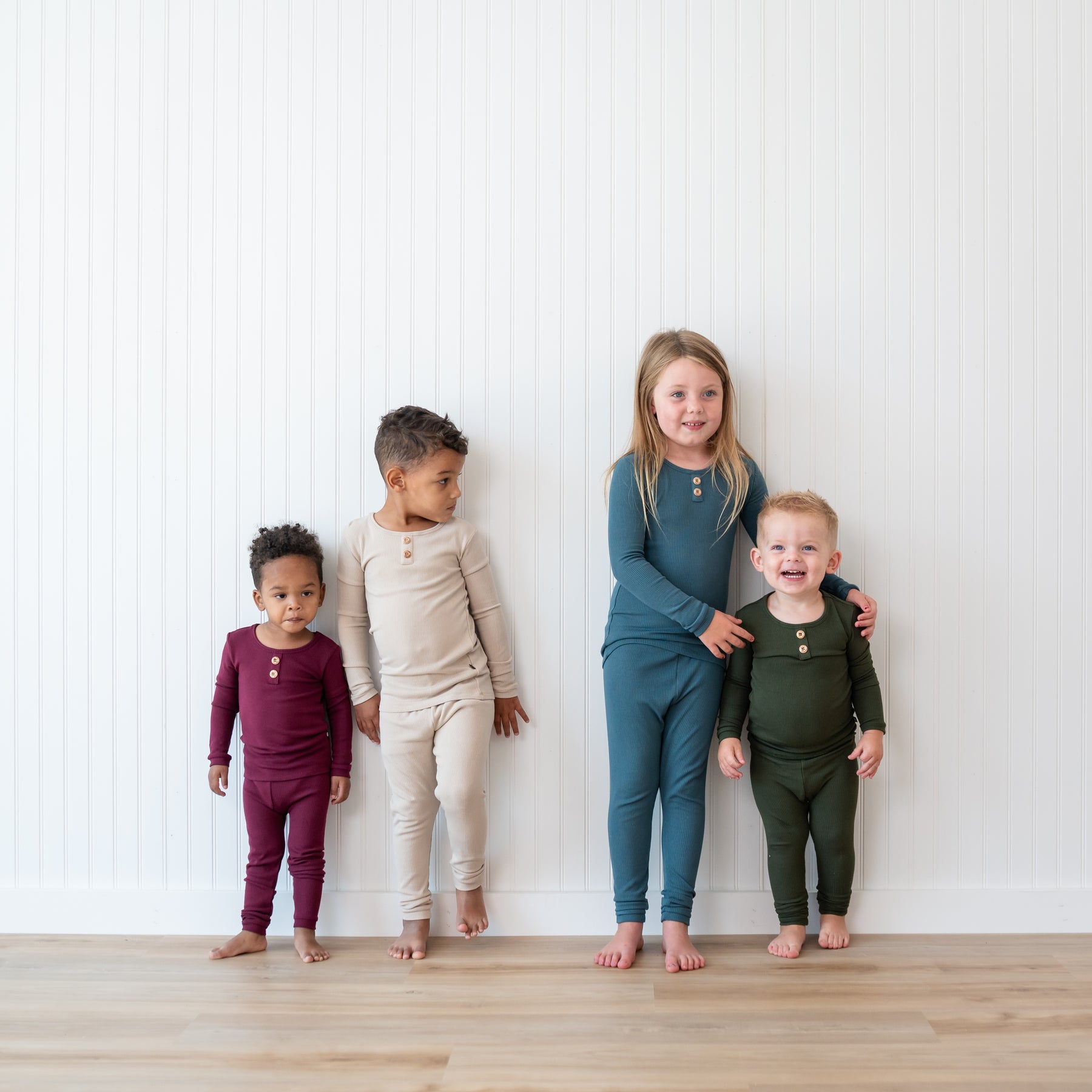 Four models standing in front of a white paneled wall wearing the Ribbed Henley Set in Fir, Atlantic, Bisque and Burgundy