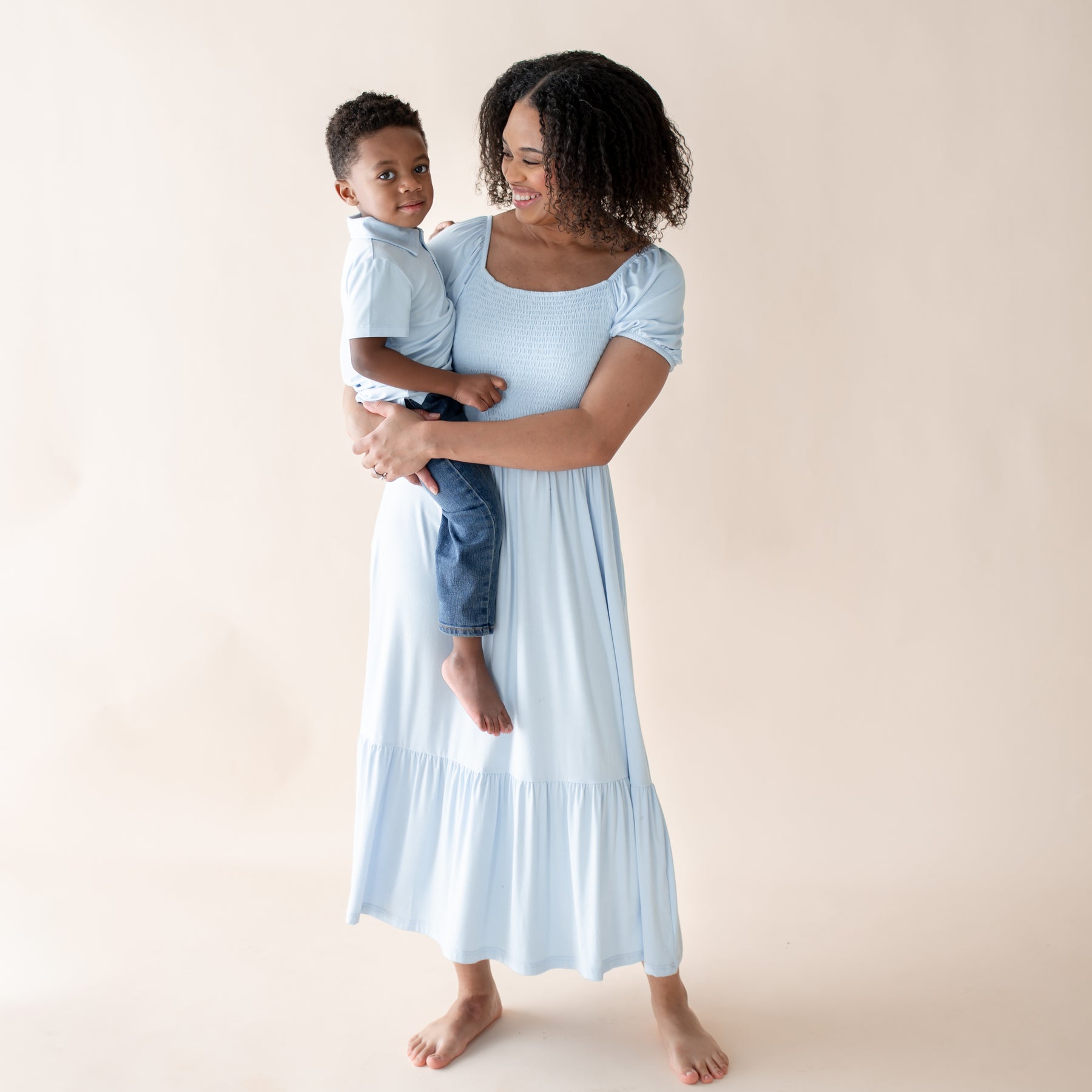 Woman in a light blue dress holding a child  in a matching blue polo against a beige background
