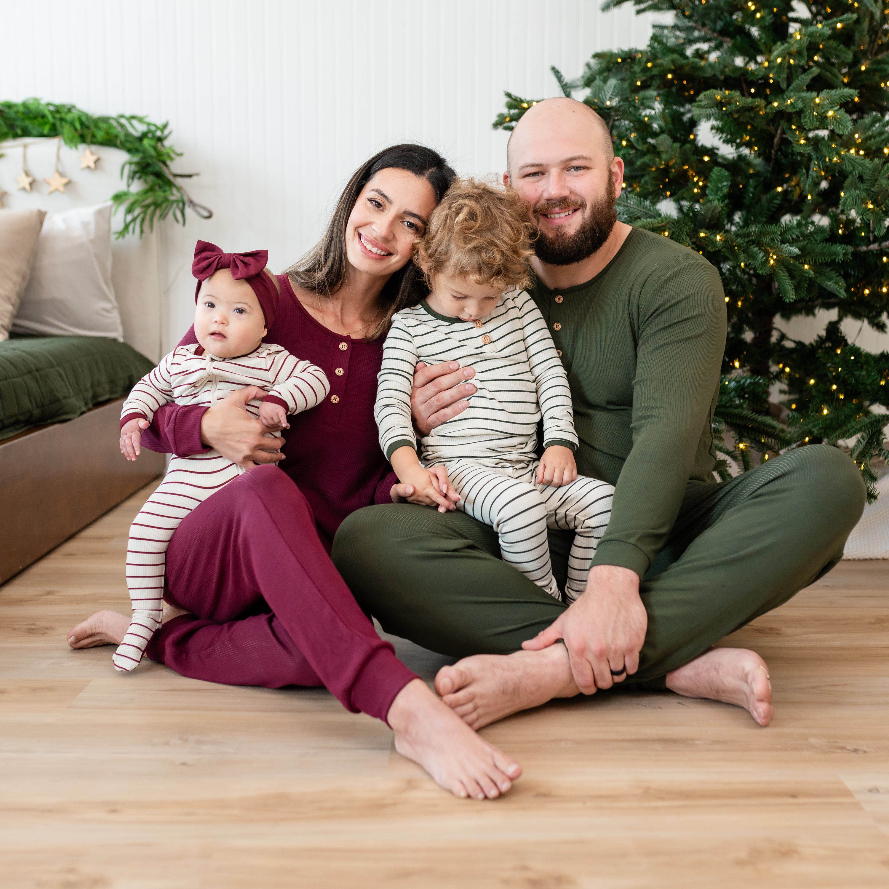 Family of four sitting on the floor beside a bed and in front of a Christmas  tree wearing the Ribbed Zipper Footie in Burgundy Stripe, Women's ribbed henley set in burgundy, Men's ribbed henley set in fir and ribbed henley set in fir stripe